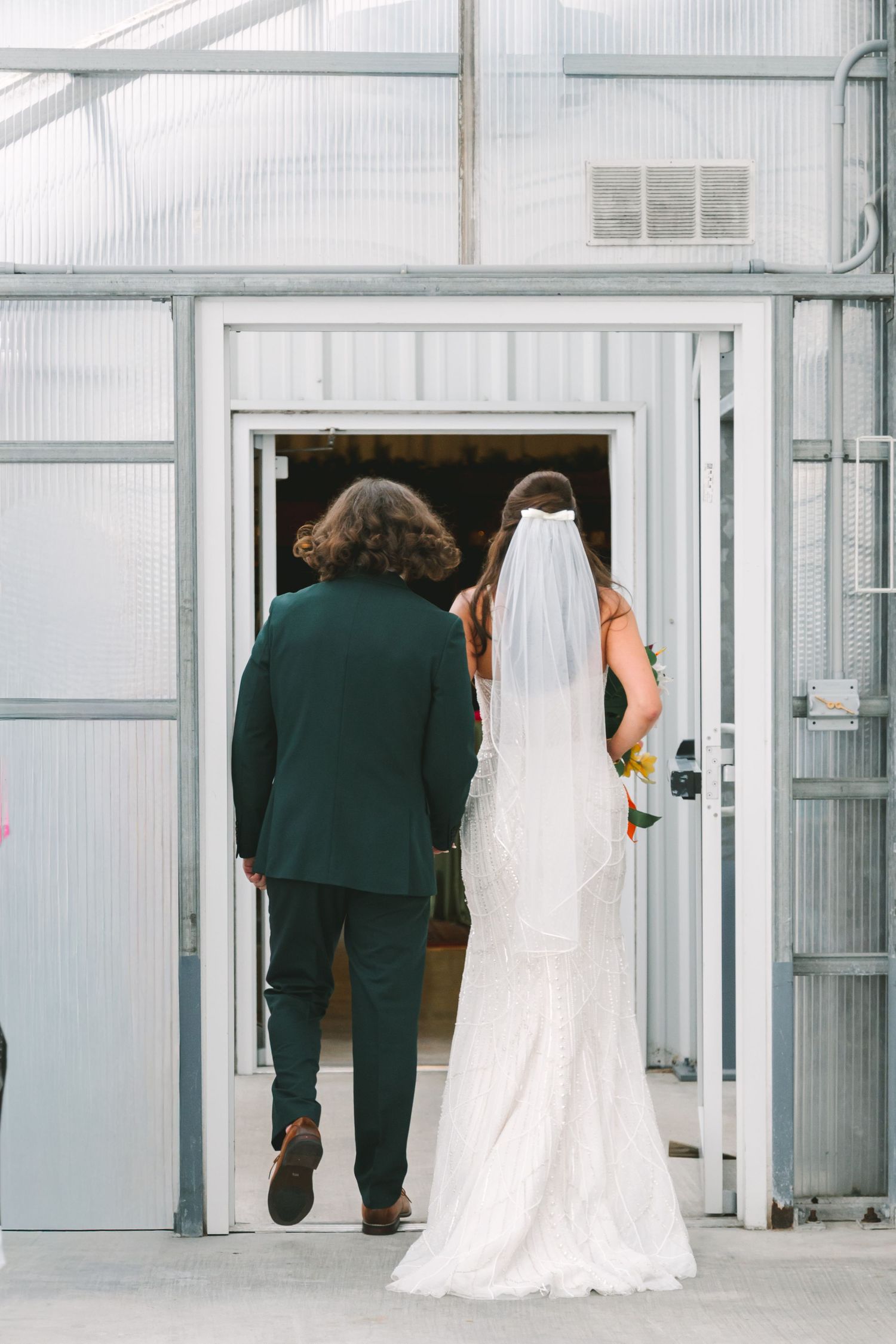 Bride in white lace gown and veil walks with groom in dark green suit through greenhouse wedding venue doors.