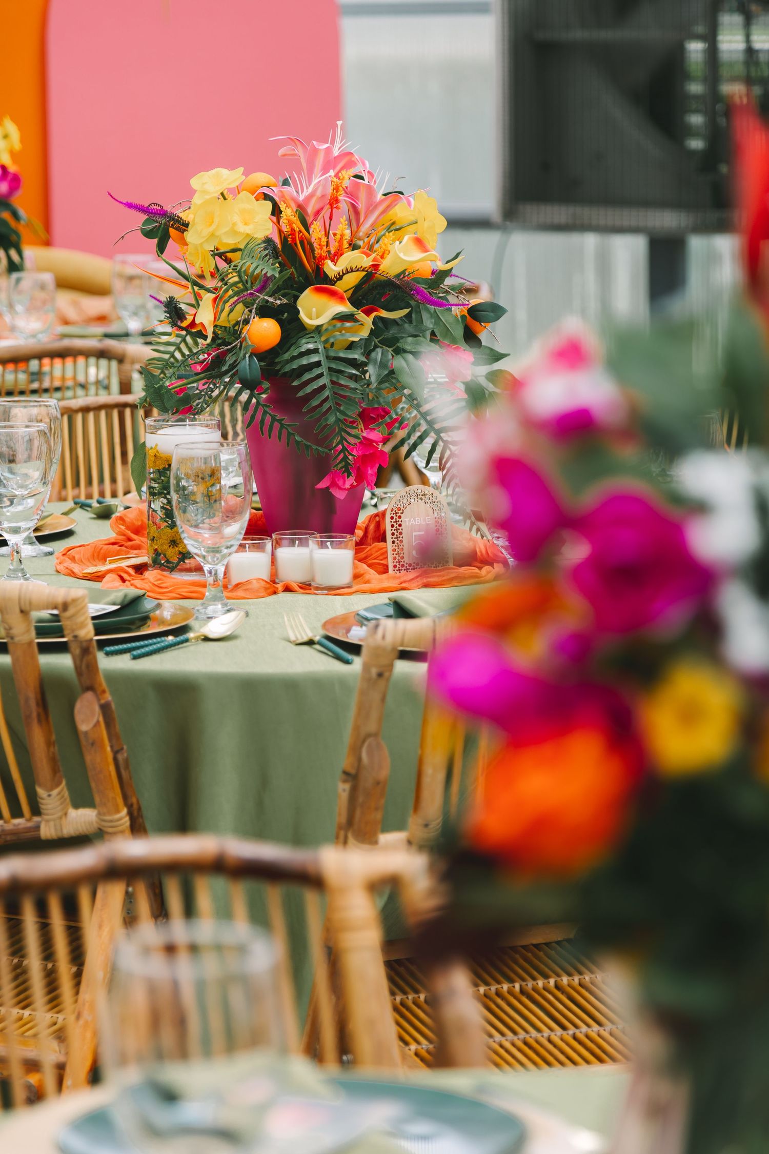 Vibrant tropical floral centerpiece on green linen table with bamboo chairs at wedding reception.