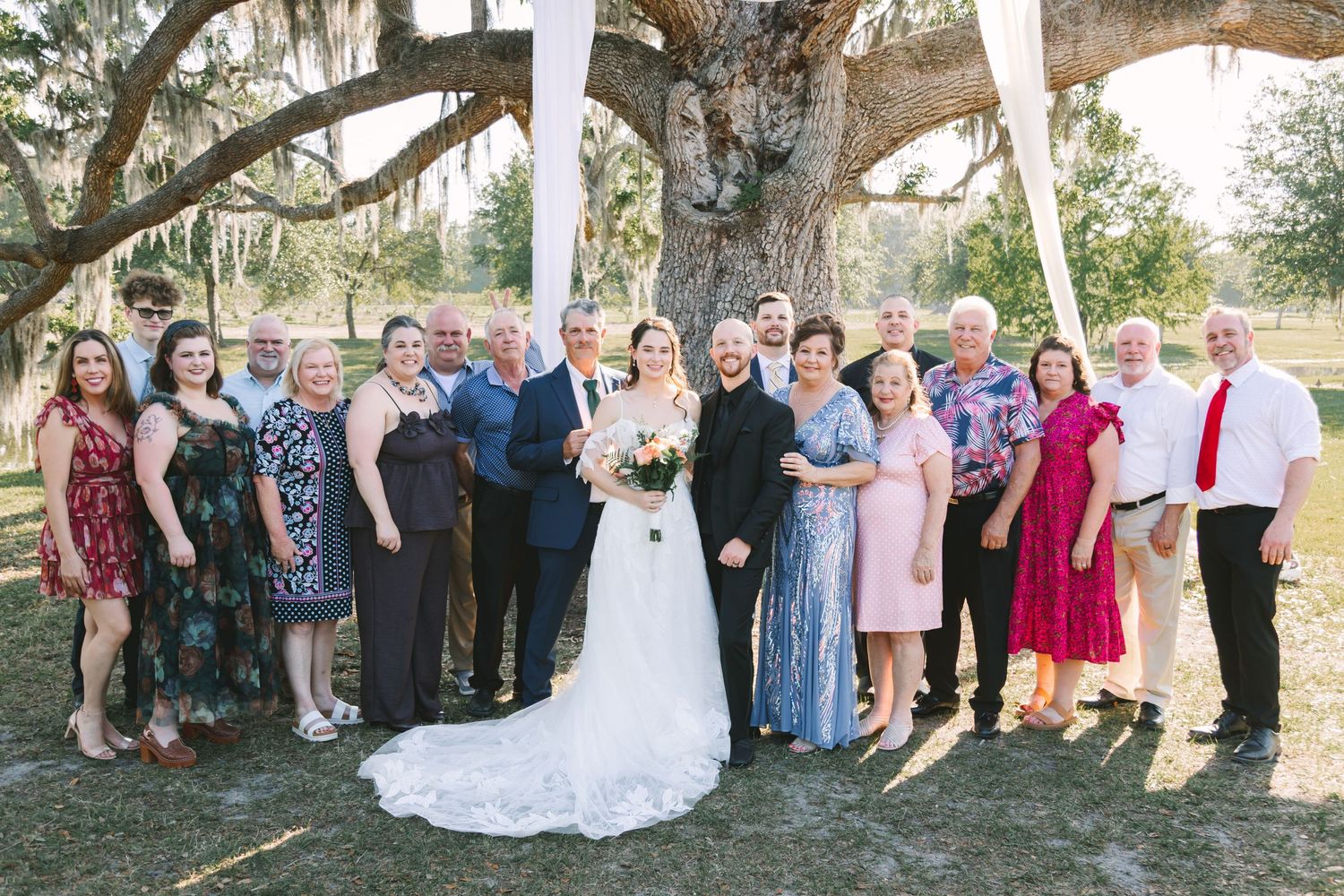Wedding party group portrait with bride and groom surrounded by family under large oak tree outdoors.