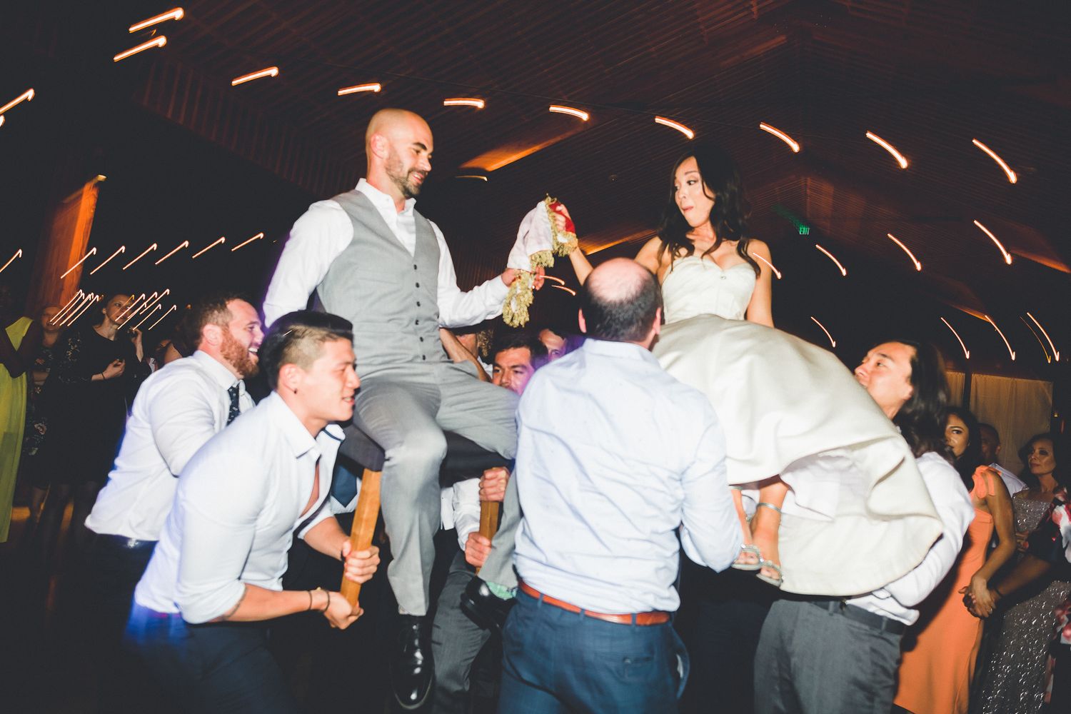 Wedding guests lifting bride and groom on chairs during a lively barn wedding reception celebration.