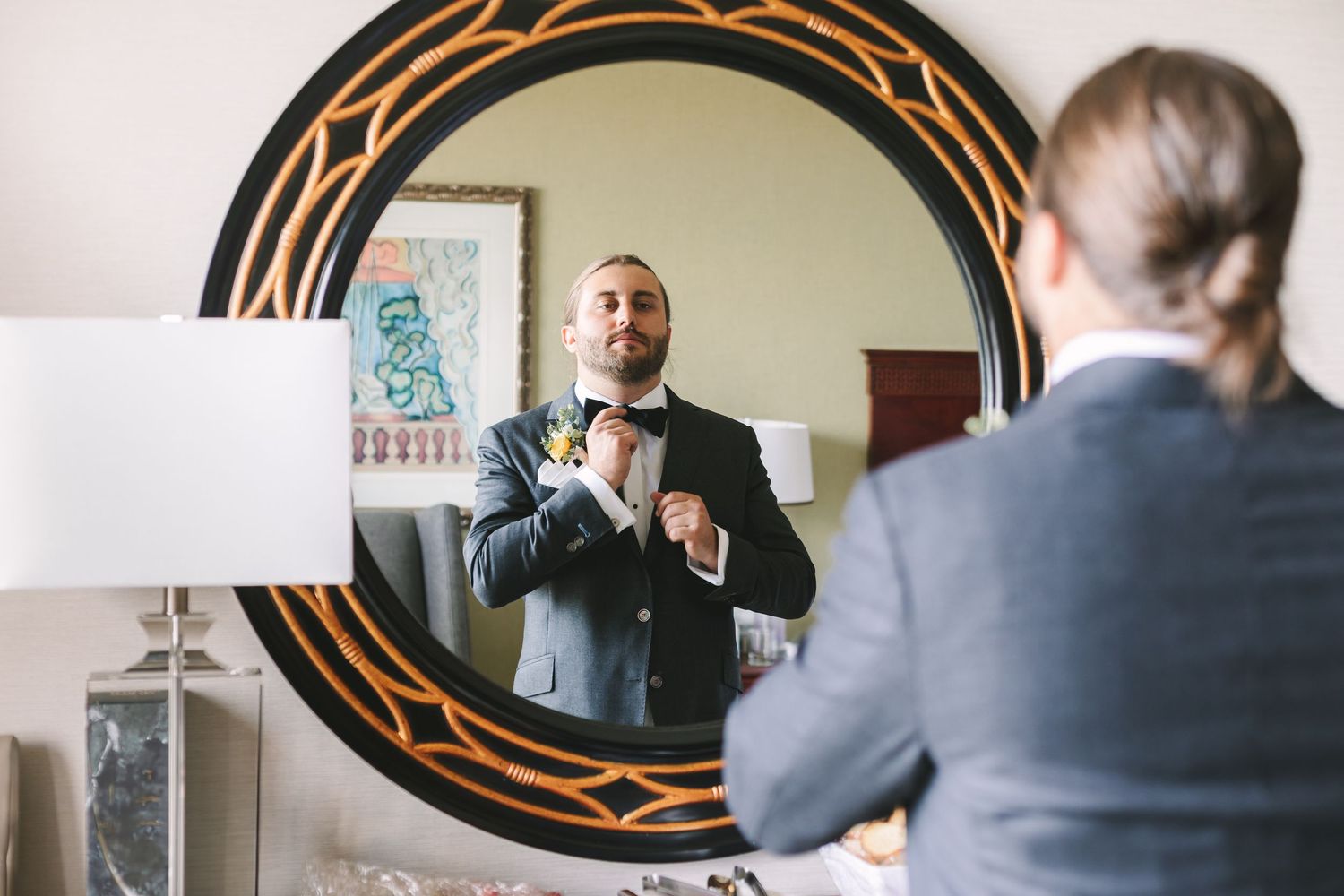 Groom adjusting his bow tie while getting ready, reflected in a decorative round black and gold mirror.