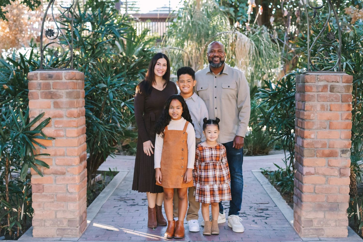 Multiracial family of five smiling together between brick pillars surrounded by lush greenery.
