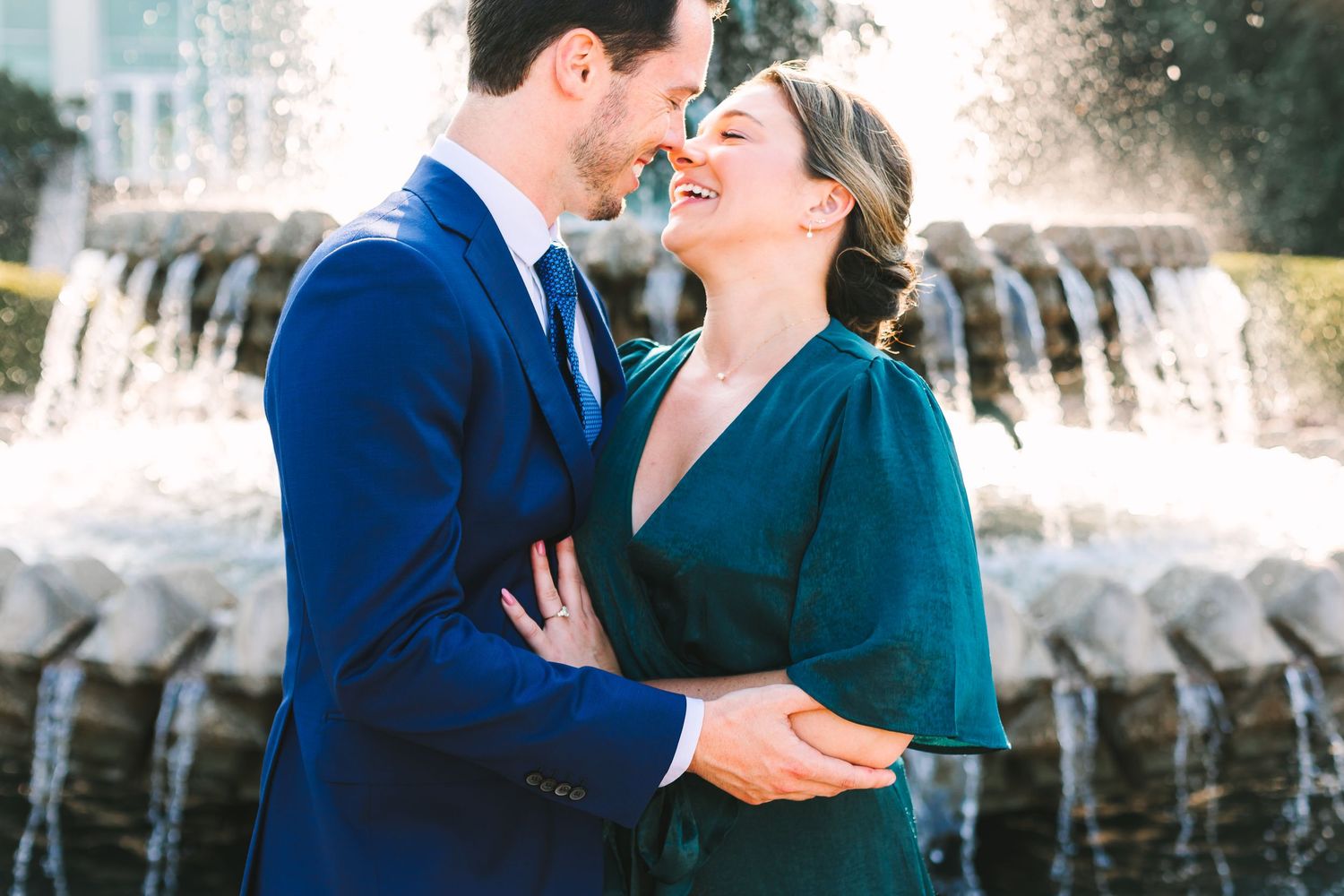 Couple laughing and embracing in front of a fountain, woman in green dress and man in blue suit.