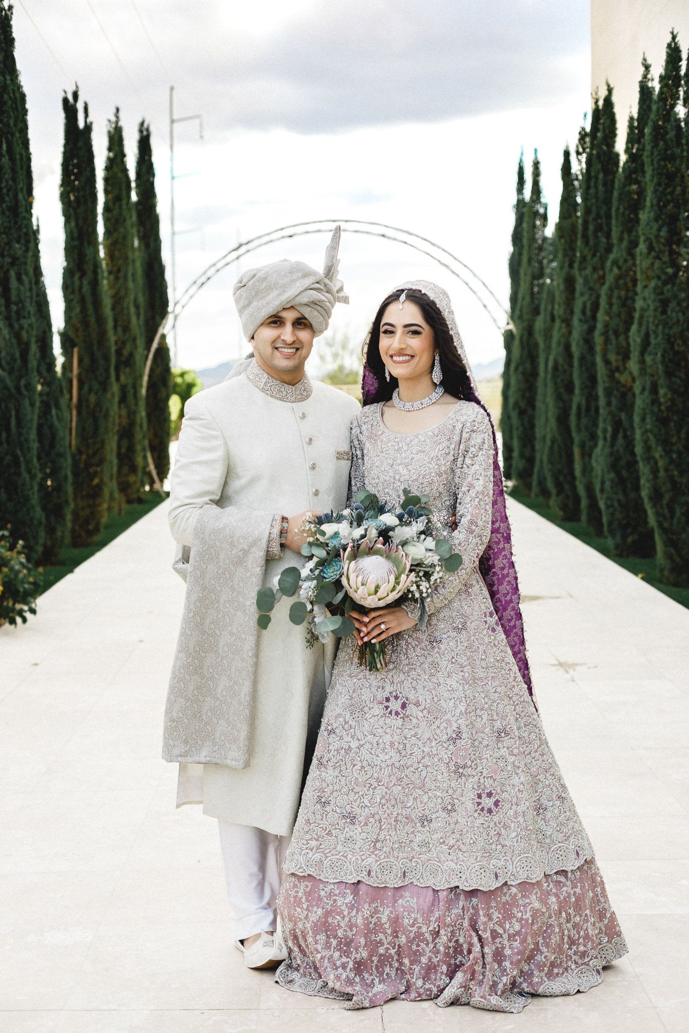 South Asian bride and groom in traditional white sherwani and silver lehenga posing outdoors with cypress trees.