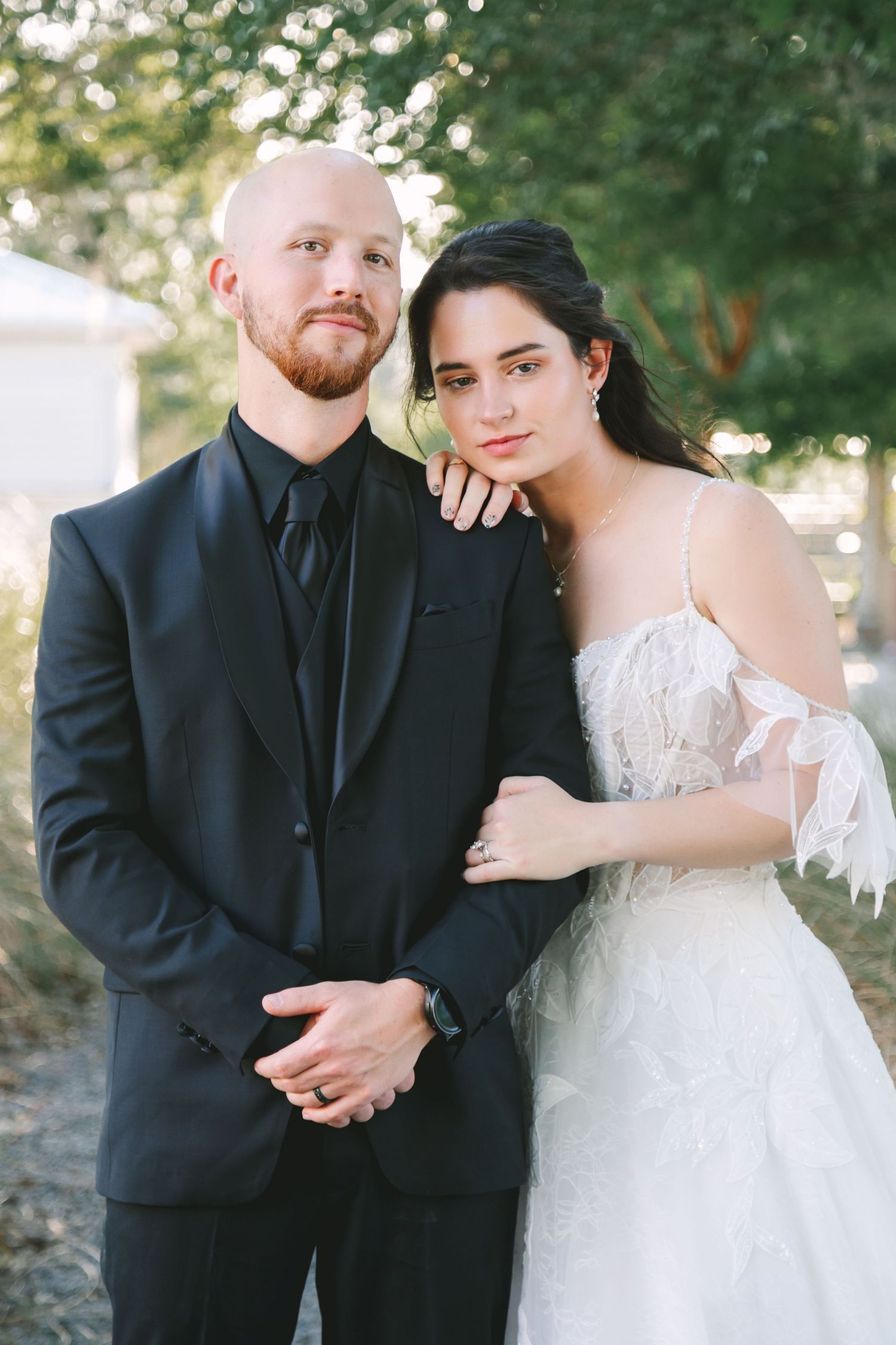 Bride in white lace gown leans on groom in black suit outdoors at elegant wedding portrait session.