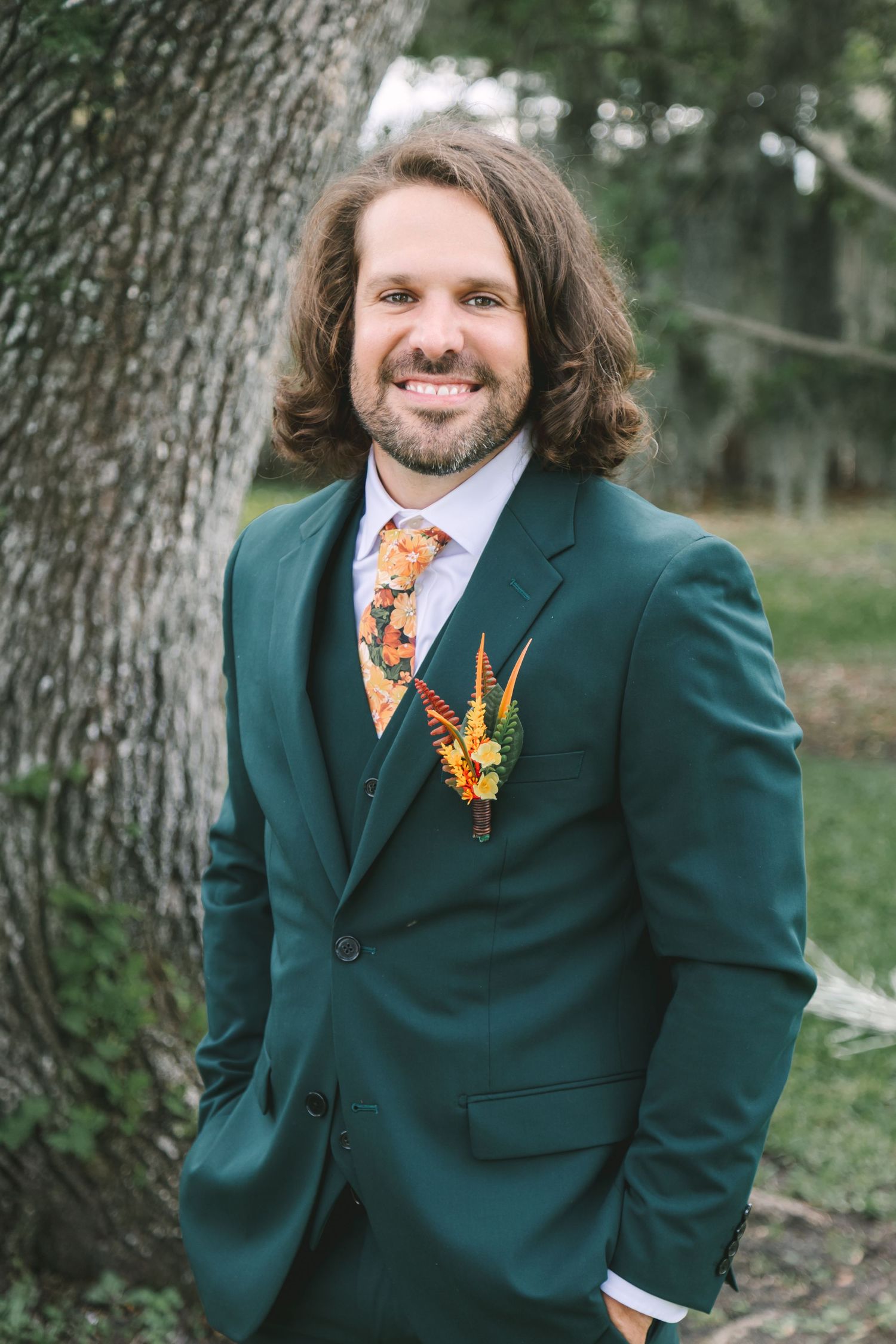 Smiling groom in green suit with floral boutonniere and patterned tie standing by tree.
