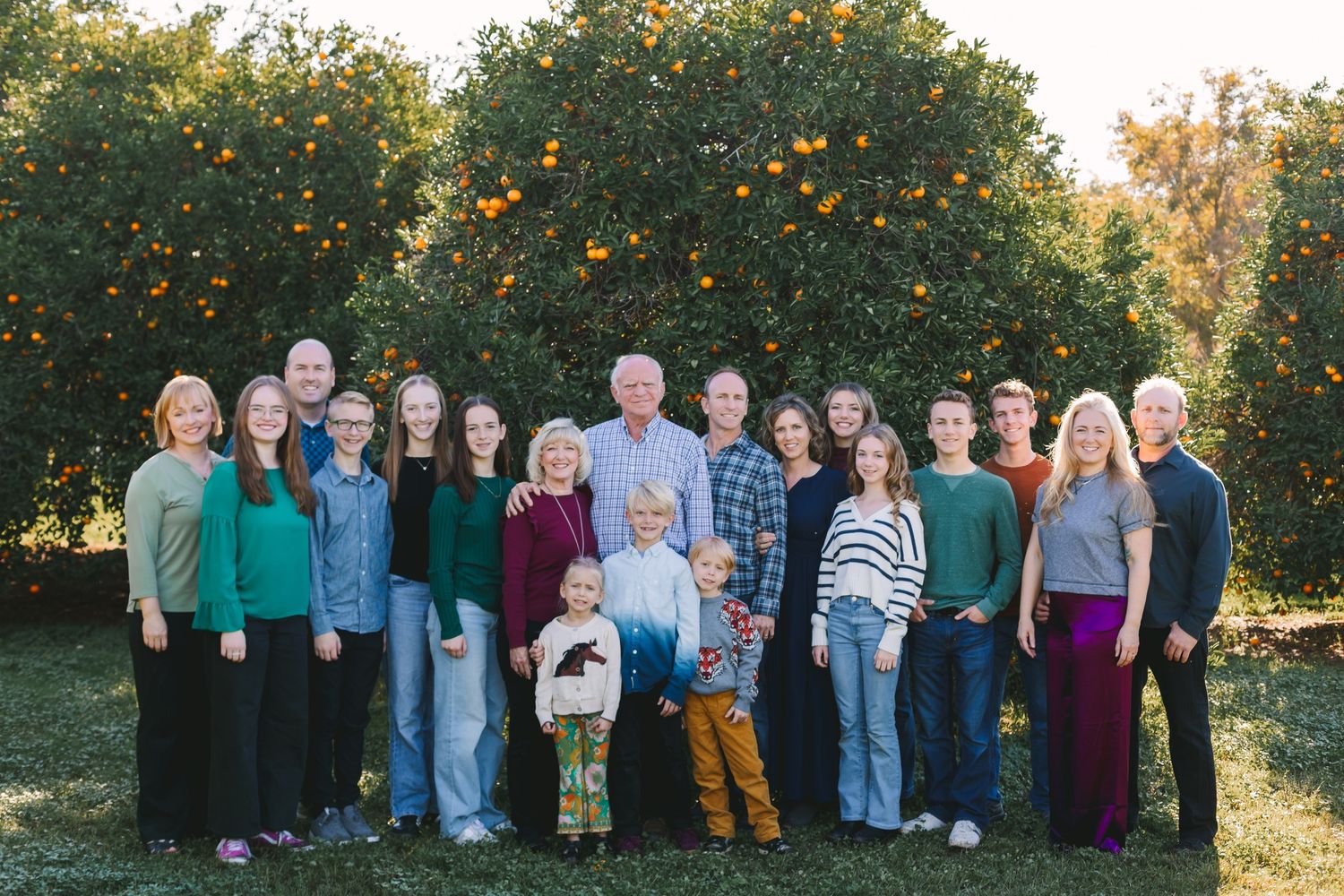 Large multigenerational family group smiling together in front of a lush orange tree orchard.