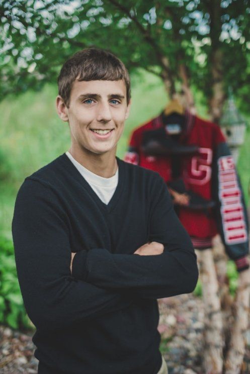 Smiling young man in black sweater with arms crossed, varsity jacket hanging in background.