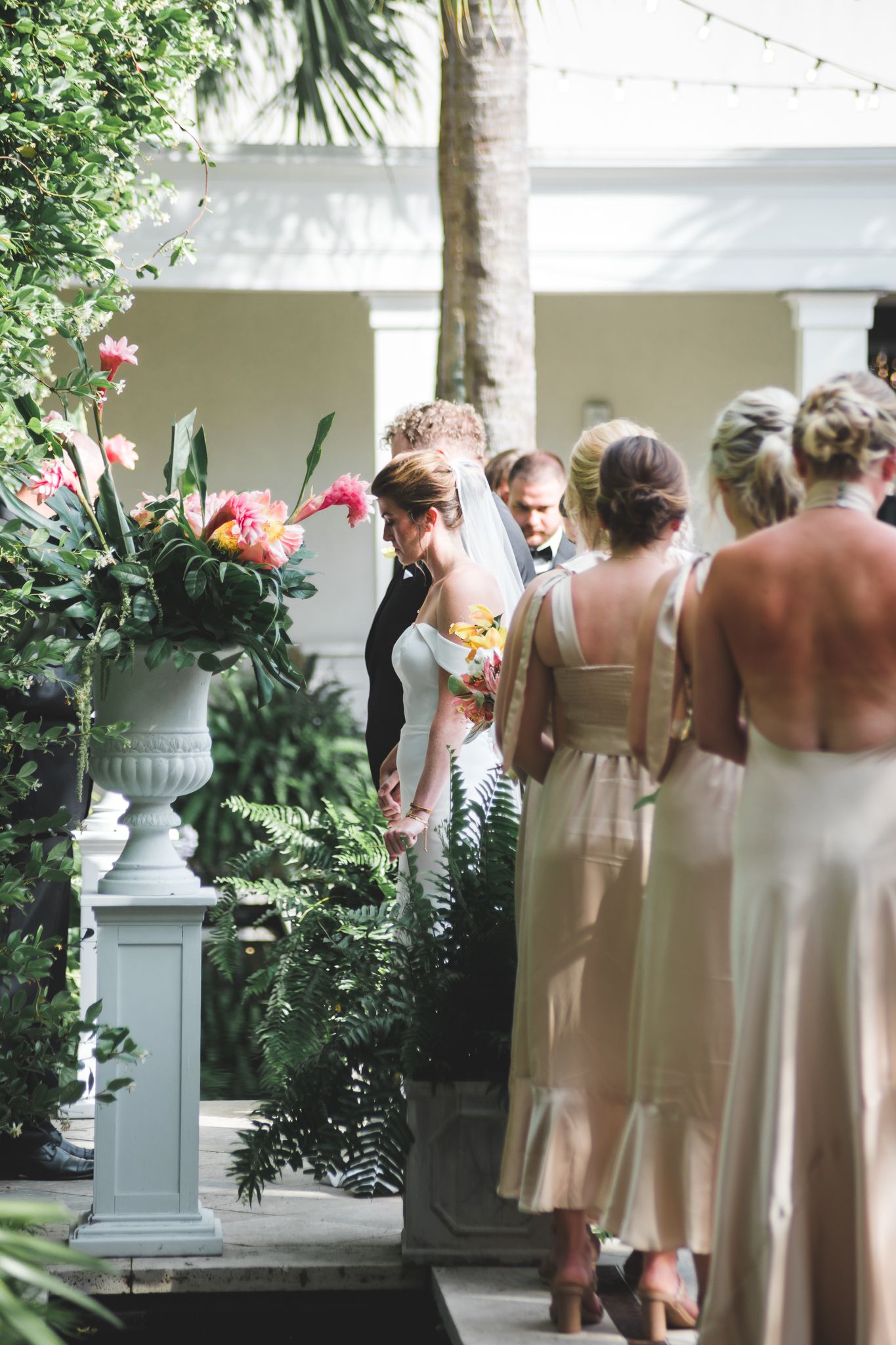 Wedding ceremony procession with bridesmaids in champagne gowns beside tropical floral arrangements.