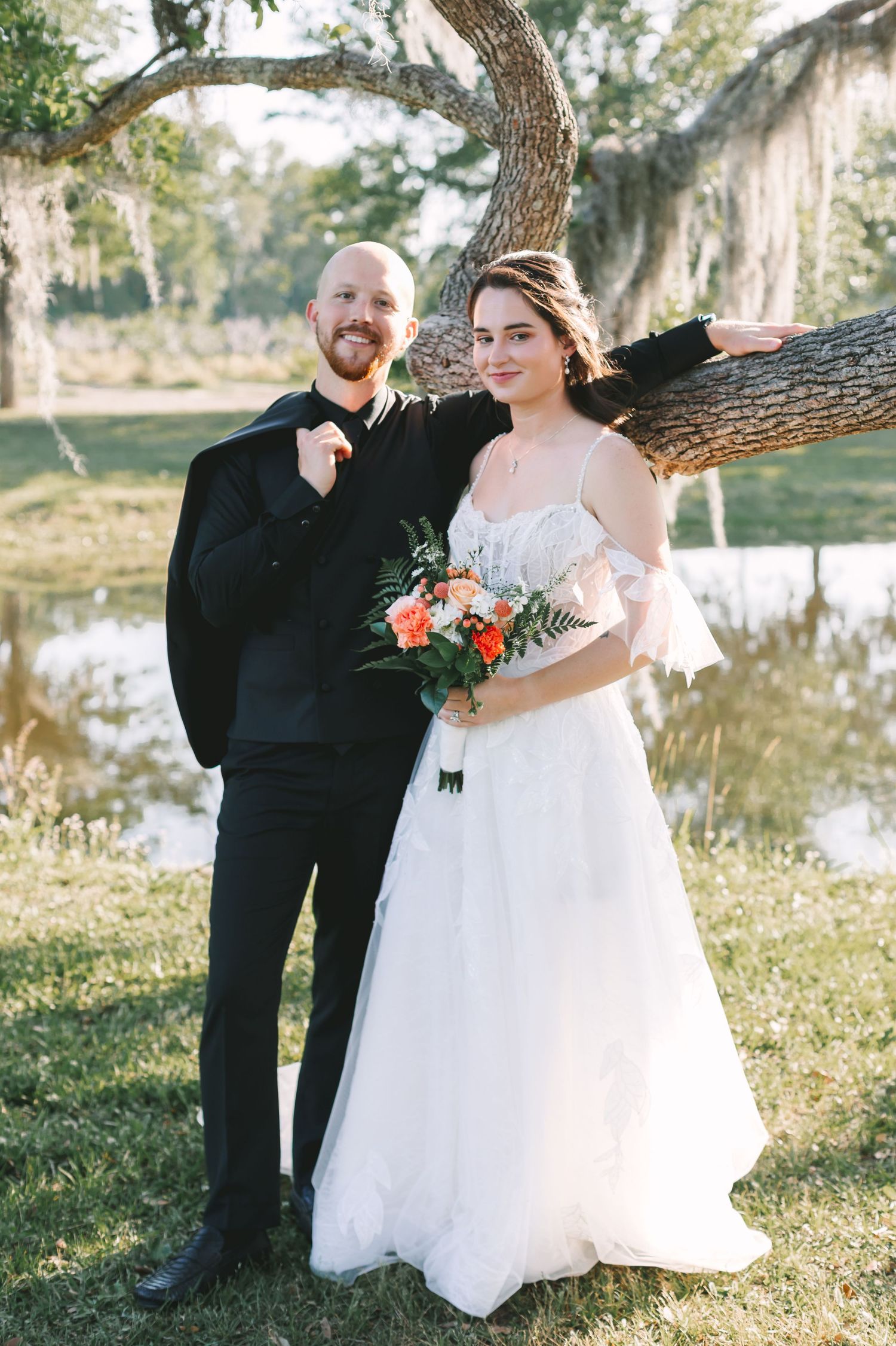 Smiling bride and groom pose together leaning against a moss-draped oak tree near a pond at Congaree and Penn.