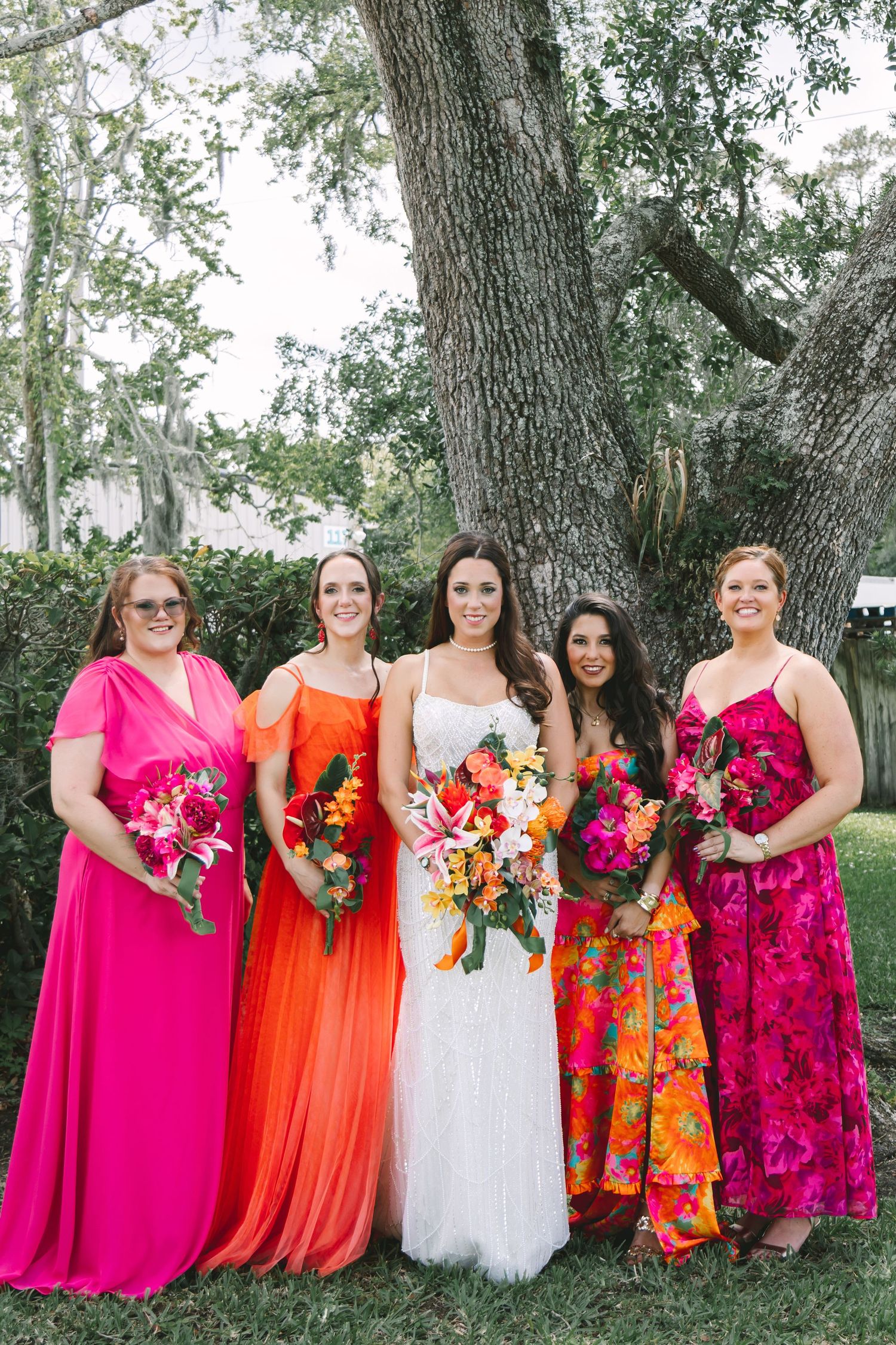 Bride in white gown with four bridesmaids in vibrant pink and orange dresses holding tropical bouquets.
