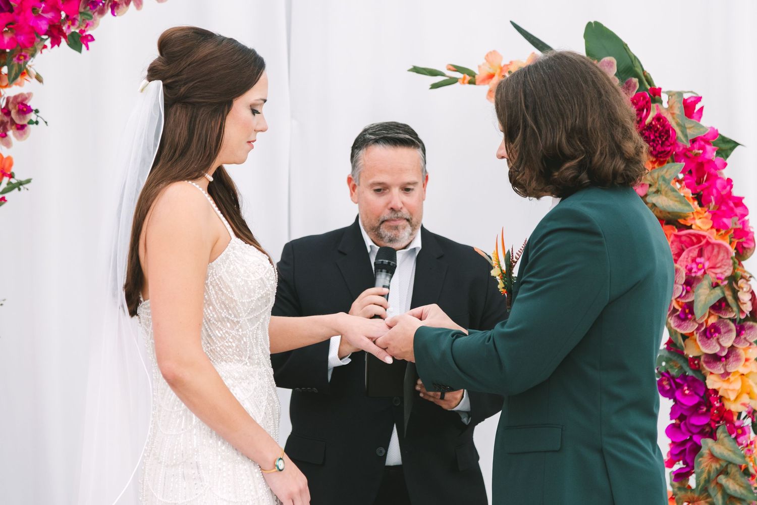 Bride and partner exchange rings at altar decorated with vibrant pink and orange tropical flowers.