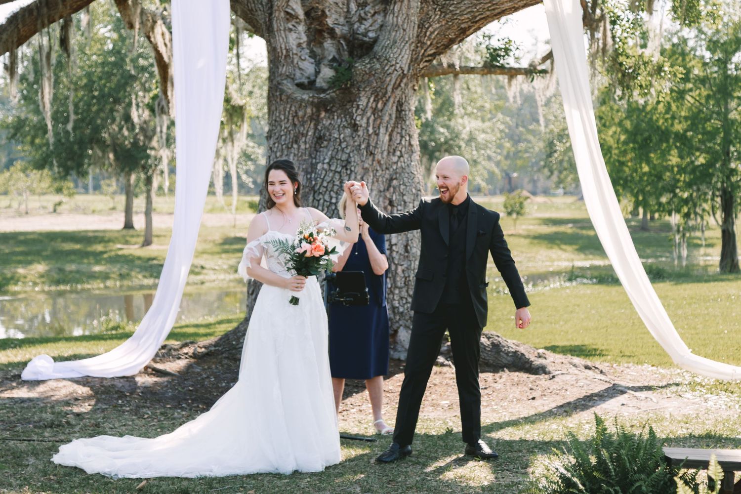 Bride and groom celebrate with officiant under oak tree with white draping at outdoor wedding ceremony.