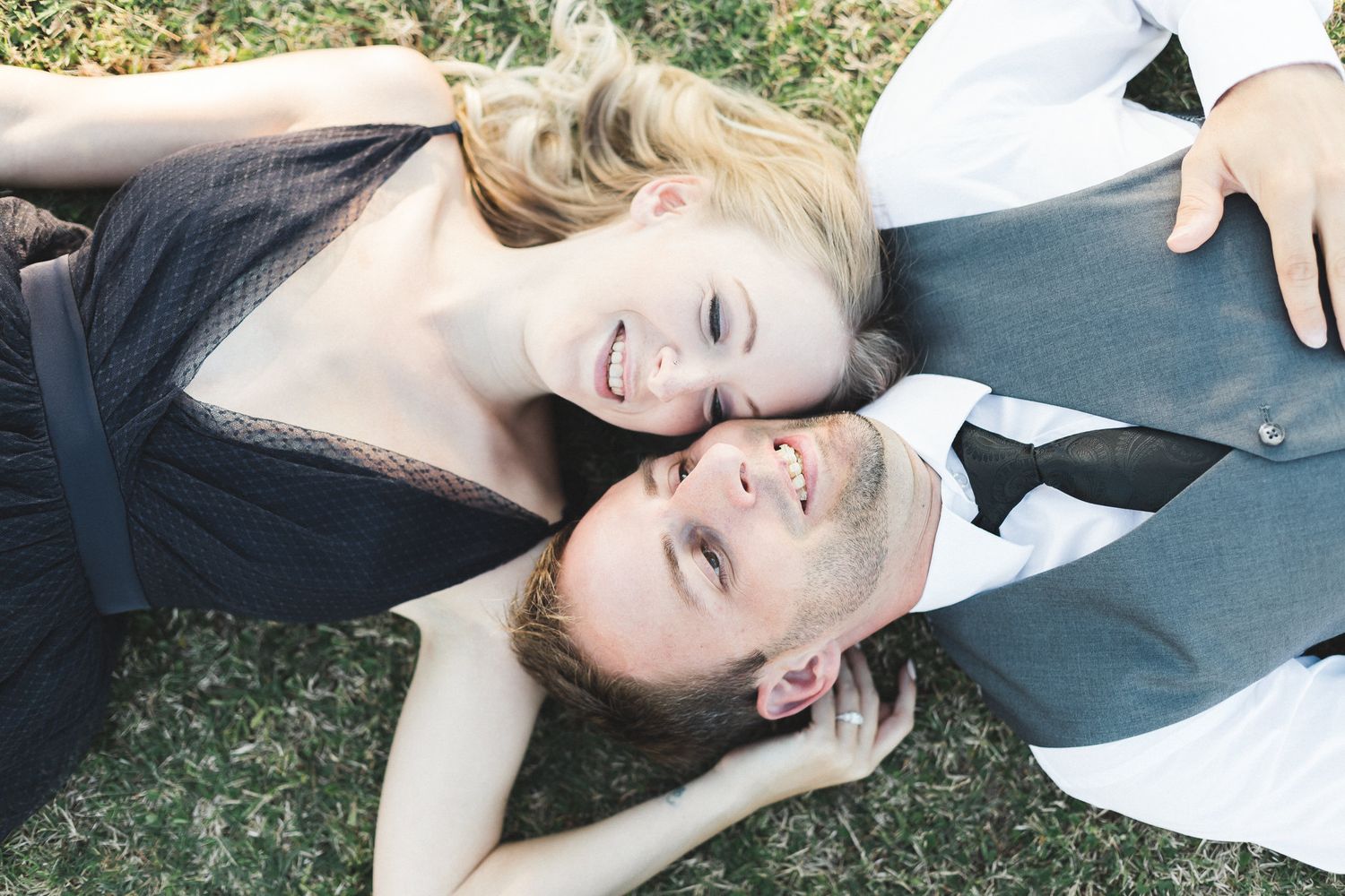 Happy couple lying on grass looking up, woman in dark dress and man in white shirt and tie smiling.