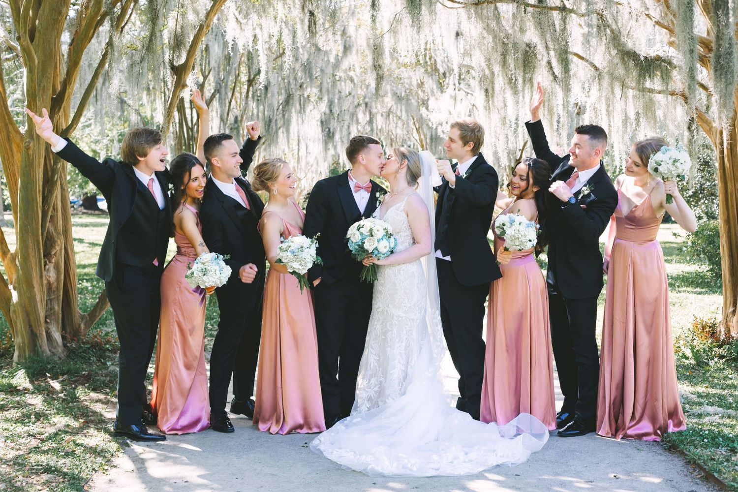 Wedding party celebrating outdoors under Spanish moss trees, bridesmaids in pink gowns and groomsmen in black tuxedos.