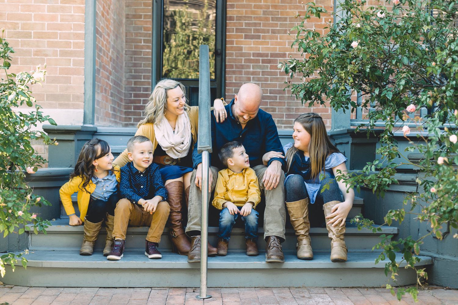 Family of six sitting and laughing together on outdoor steps surrounded by lush green plants.