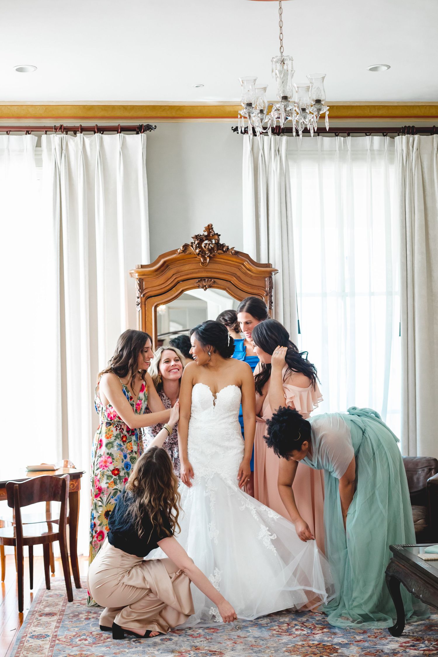 Bride in white gown surrounded by bridesmaids helping her get ready in an elegant chandelier-lit room.