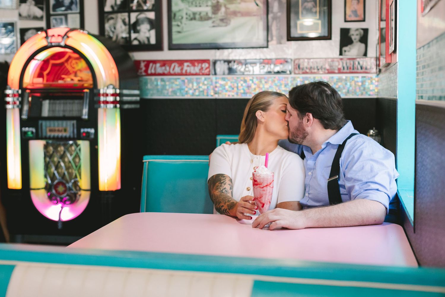 Couple kissing in a retro diner booth beside a colorful jukebox, sharing a pink milkshake.