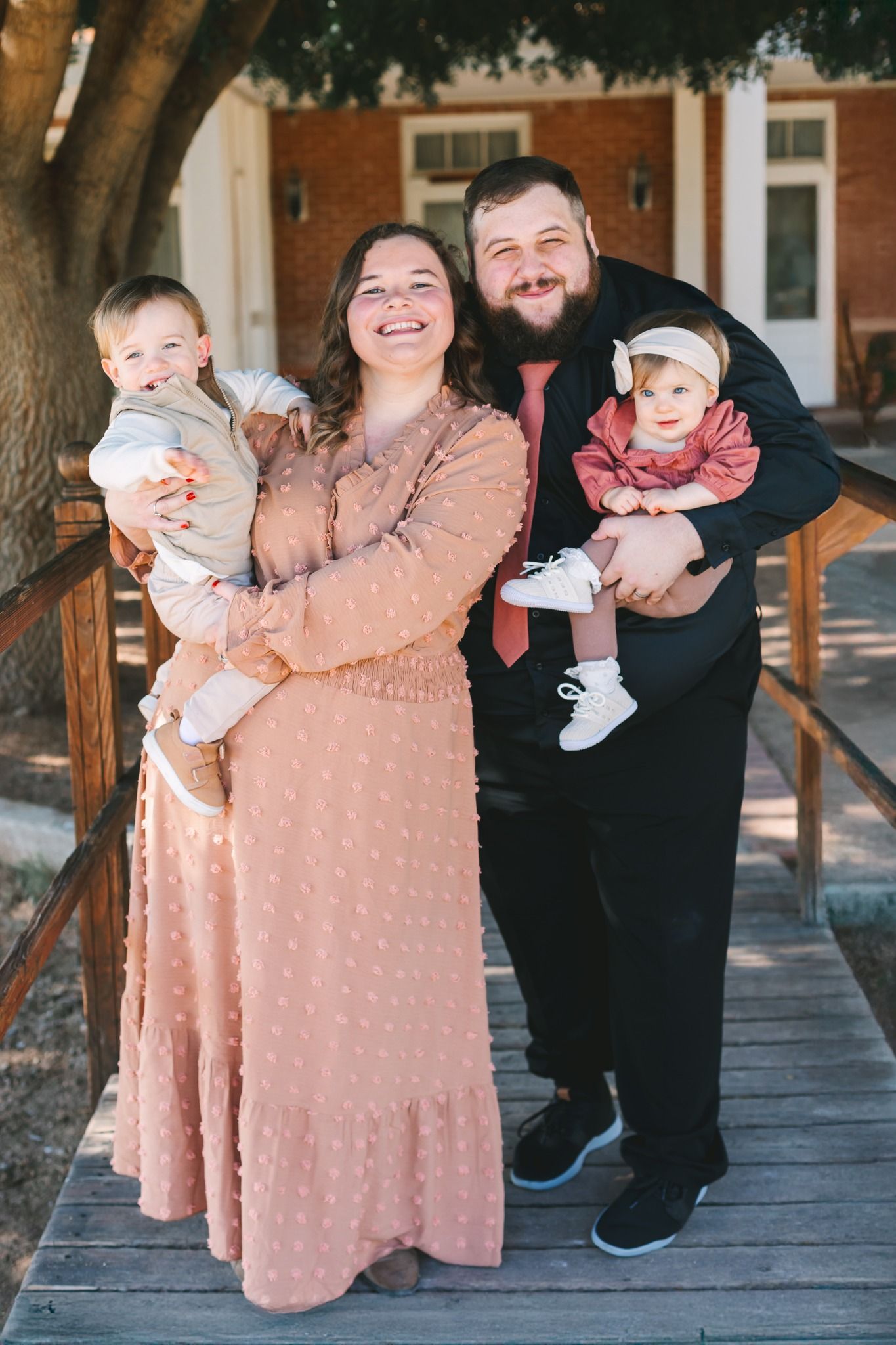 Young couple holding two toddlers smile together on wooden bridge near brick building.