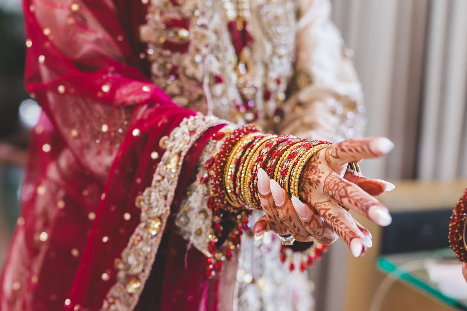 Close-up of South Asian bride\u2019s hands adorned with intricate henna, gold bangles, and red bridal attire.