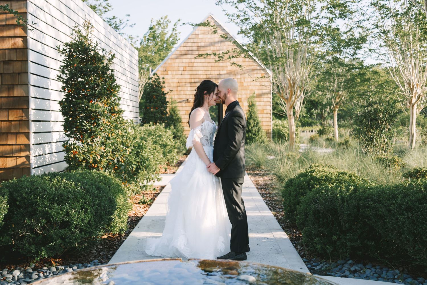 Bride and groom share romantic moment on garden pathway surrounded by manicured hedges and greenery.