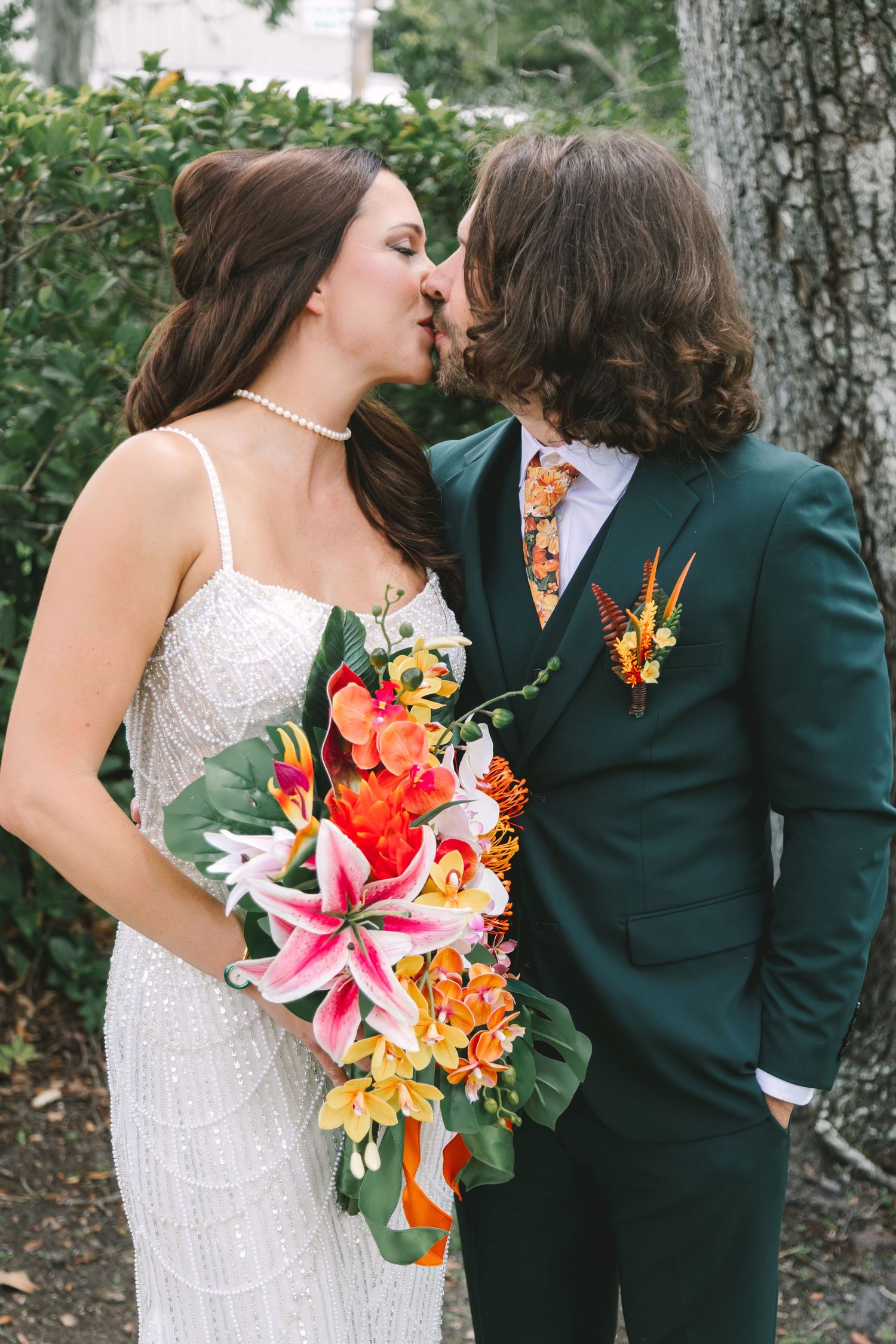 Bride and groom share a kiss outdoors, she holds a tropical bouquet, he wears a green suit.