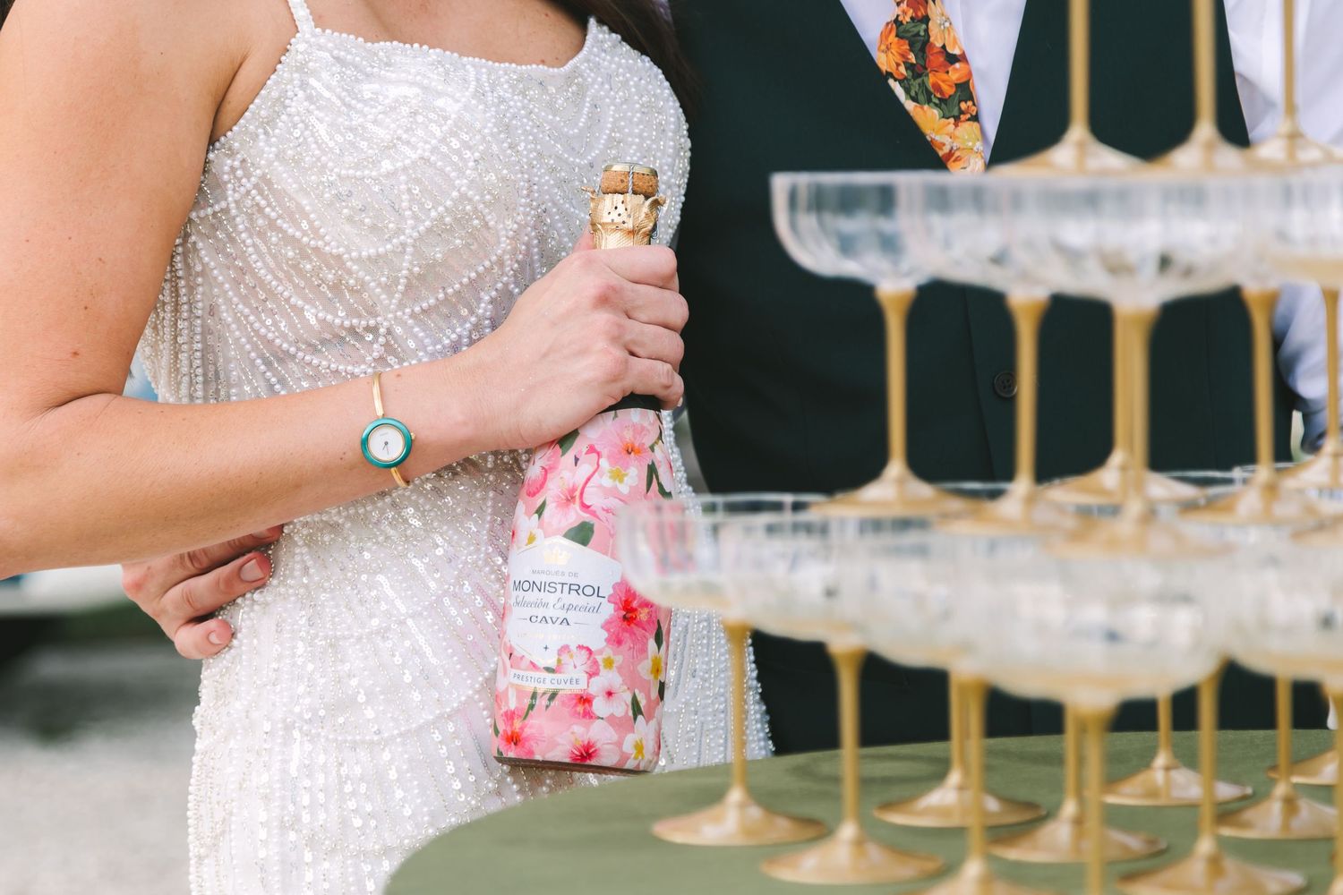 Bride in beaded gown holding floral champagne bottle near gold champagne tower at reception.