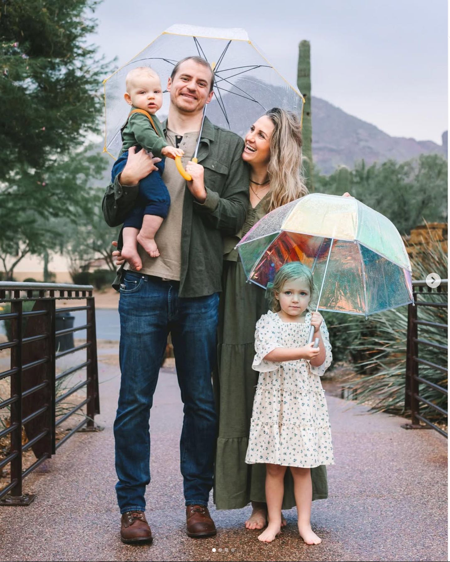 Family of four posing with clear umbrellas on a rainy day outdoors, parents holding young children in a scenic setting.