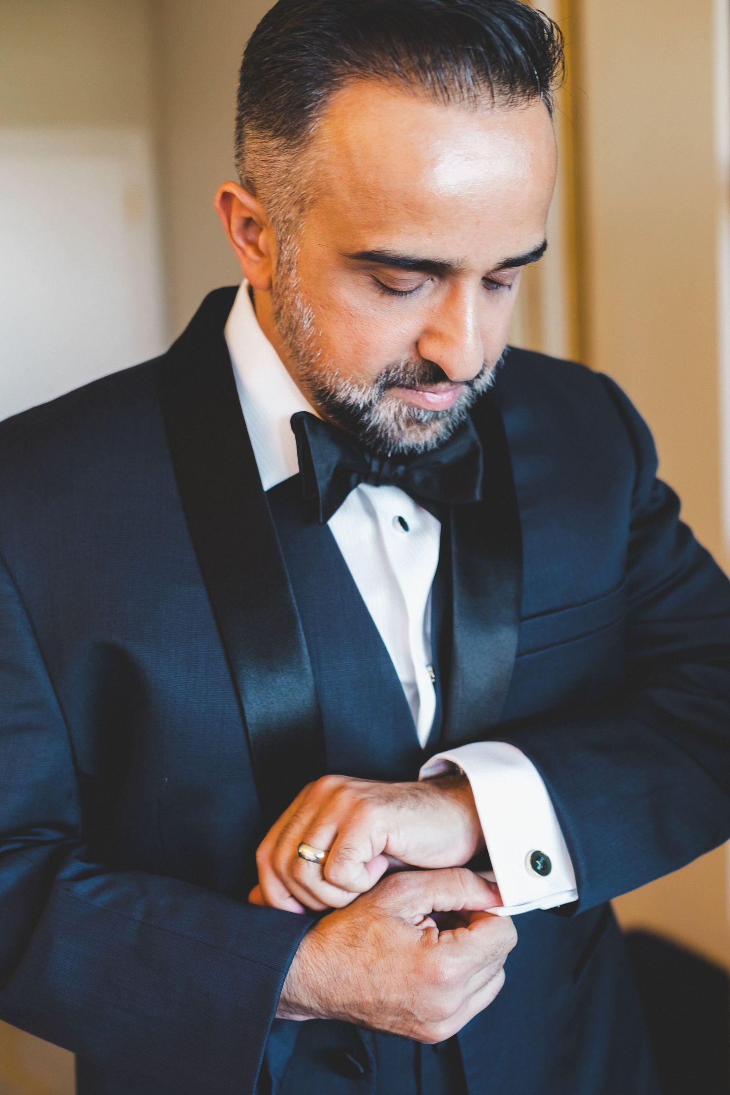 Groom in black tuxedo with bow tie adjusting cufflinks while getting ready for wedding ceremony.
