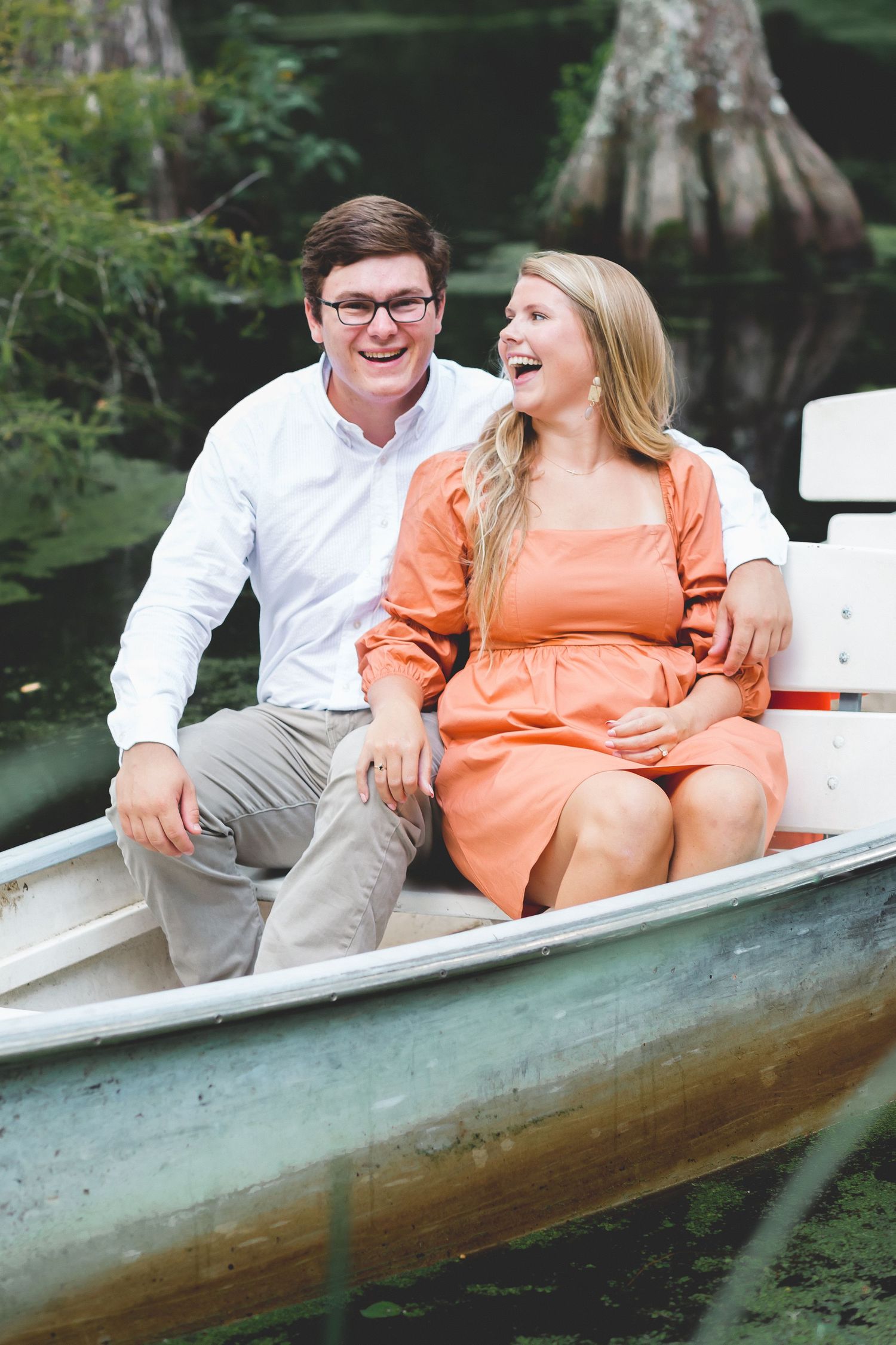 Laughing couple sitting together in a rowboat surrounded by lush green trees.