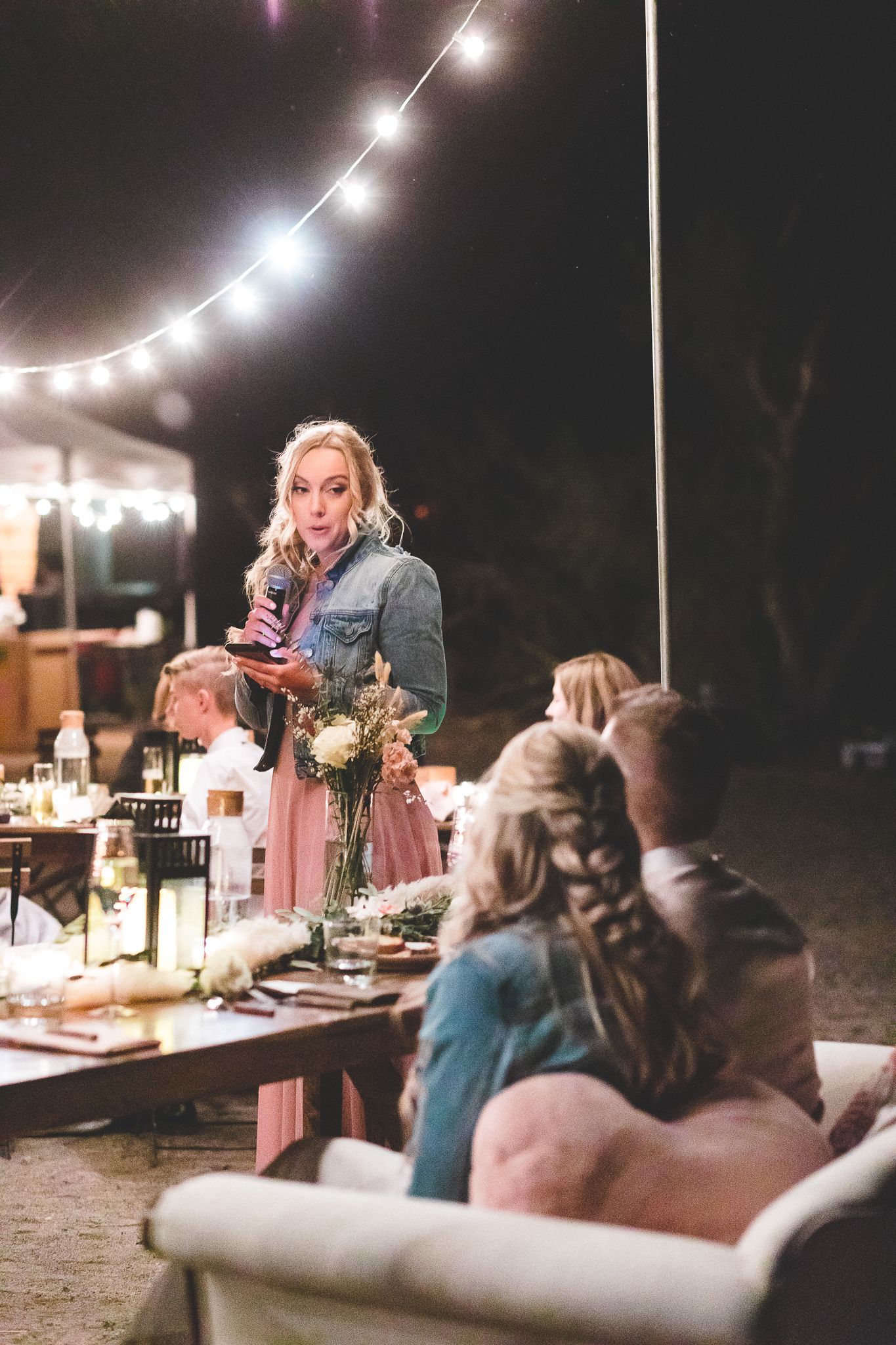 Blonde woman giving a heartfelt toast at an outdoor wedding reception under string lights.