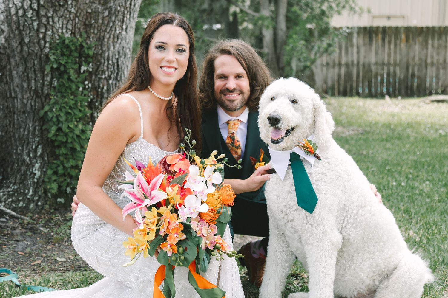 Bride and groom smiling with large white fluffy dog wearing green tie outdoors at wedding.