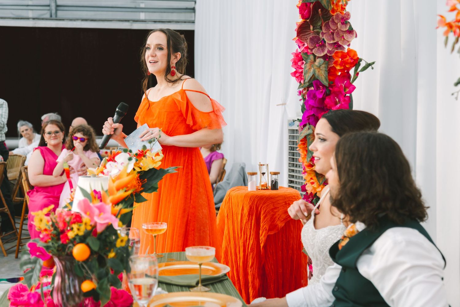 Woman in orange dress giving a speech at a colorful wedding reception with bright floral arrangements.
