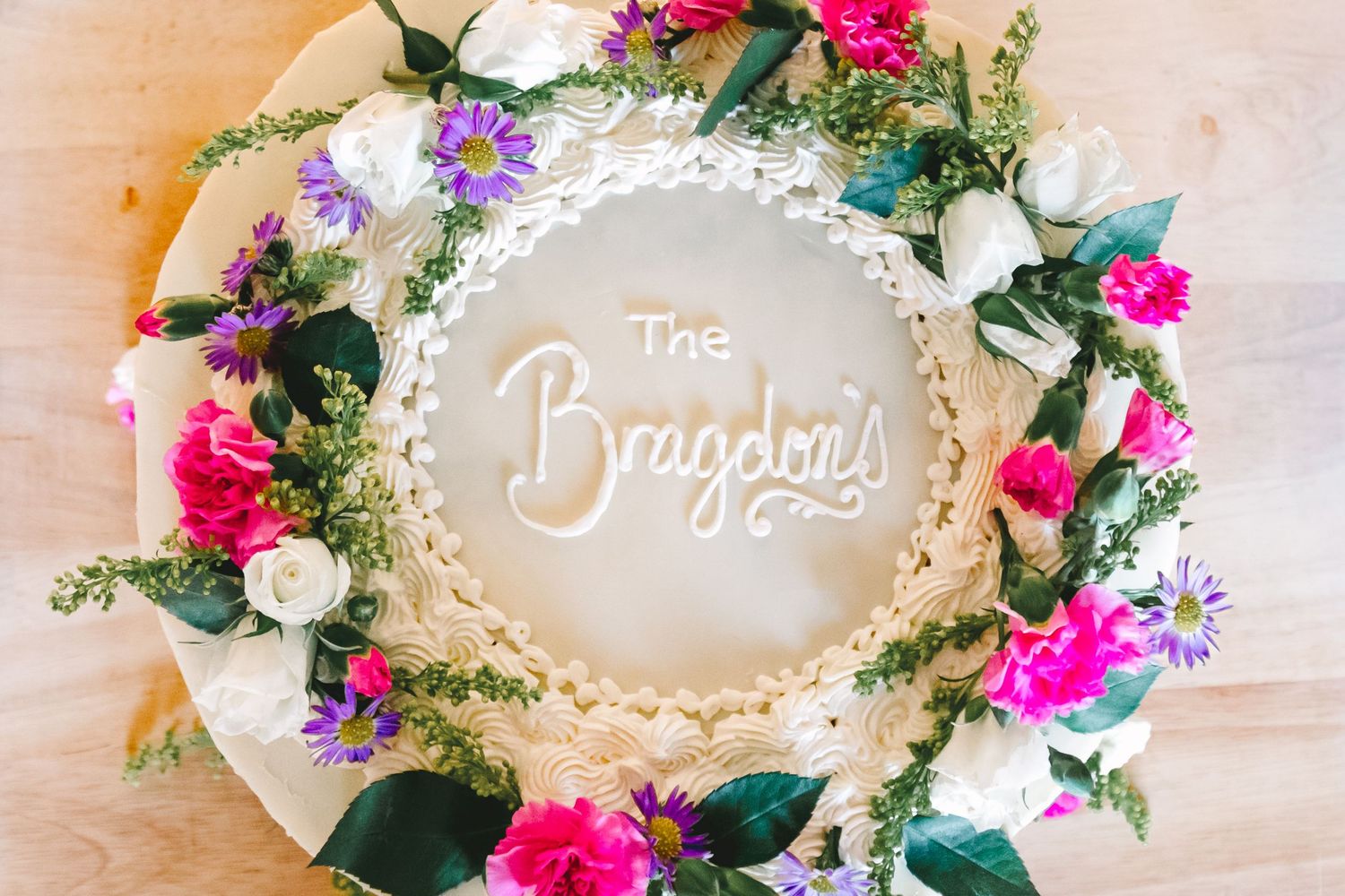 Overhead view of round wedding cake decorated with colorful flowers reading The Bragdons in script.