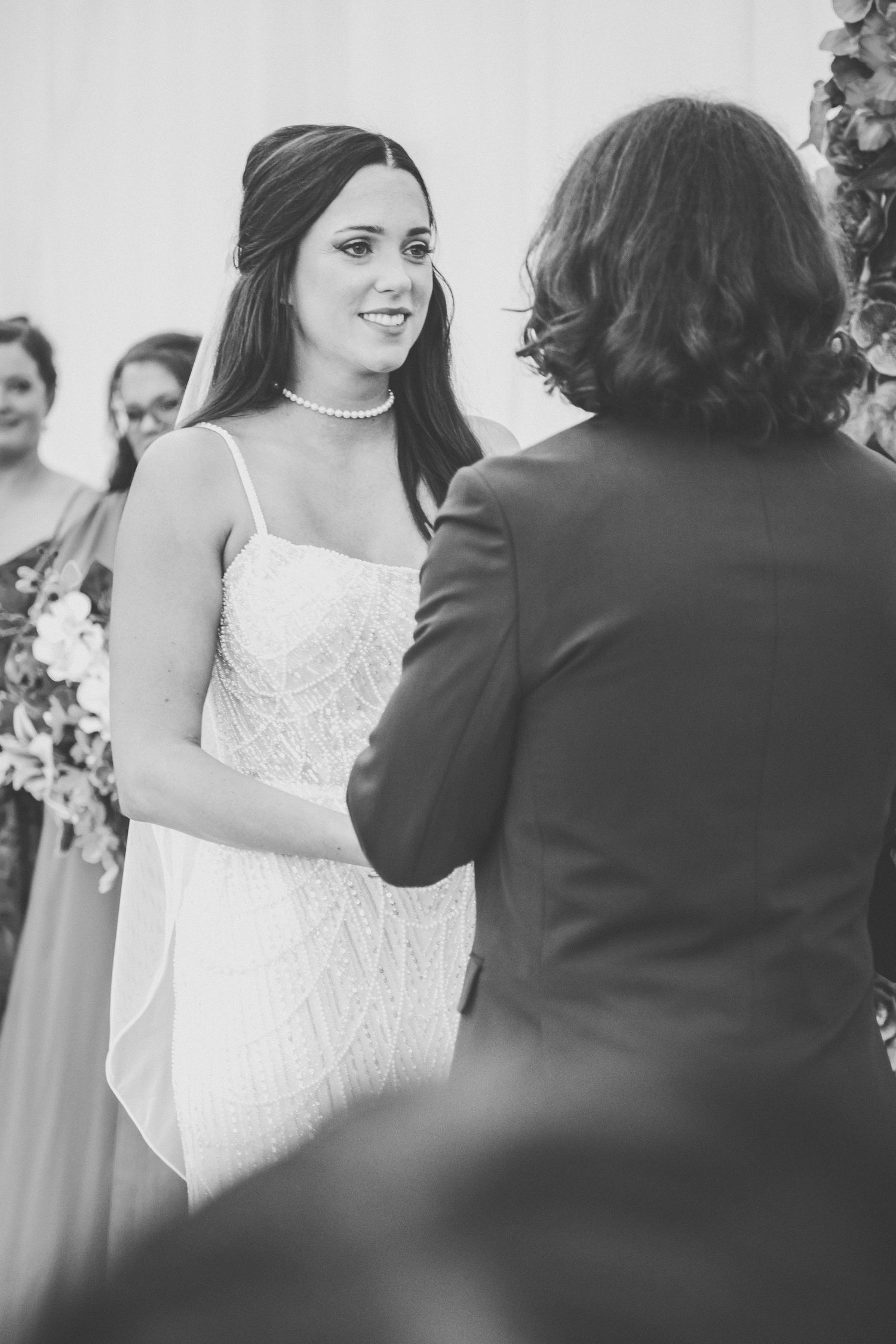 Black and white photo of smiling bride in pearl necklace exchanging vows at wedding ceremony.