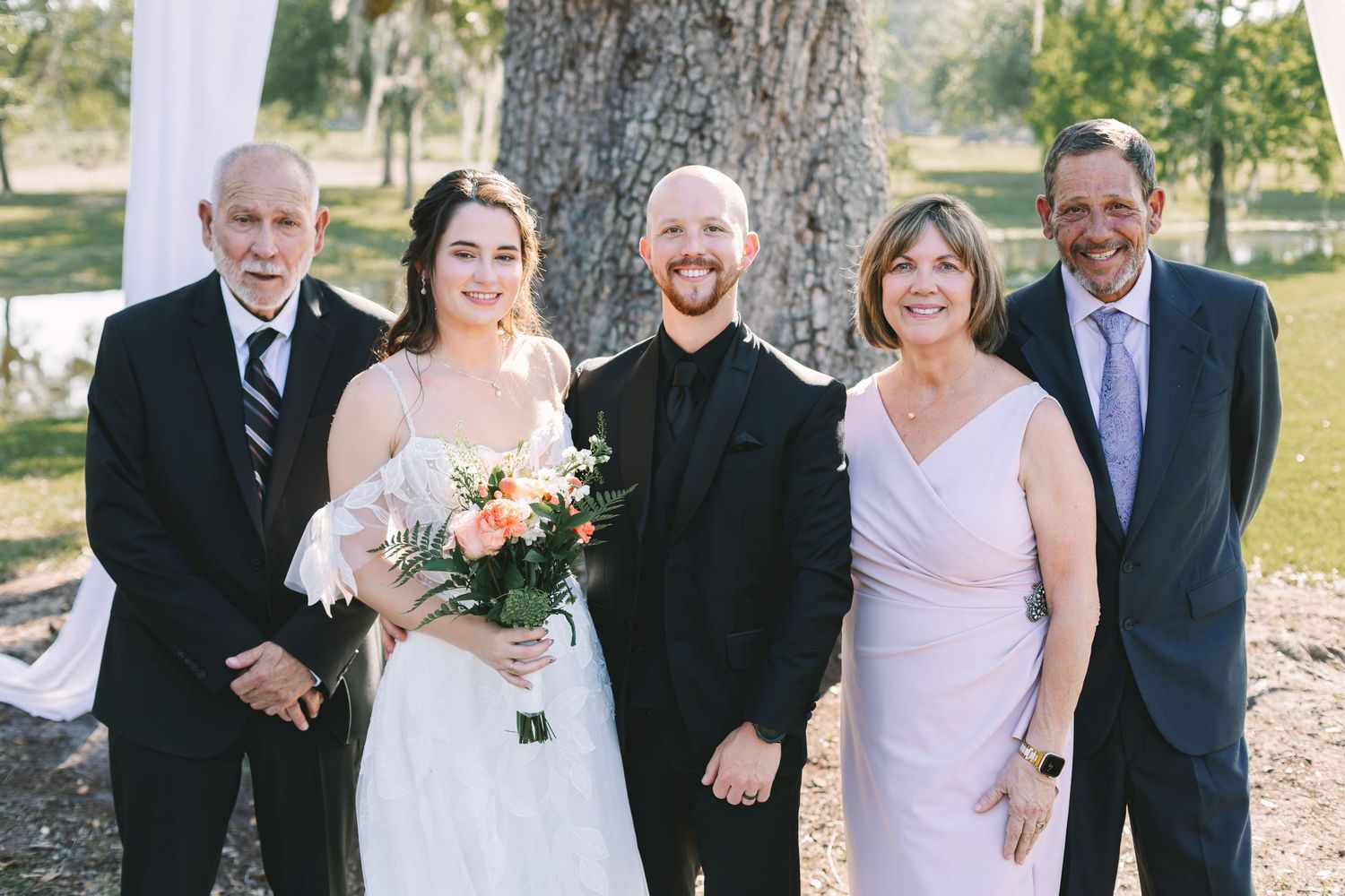 Wedding party of five posing outdoors near a large tree, bride holding a colorful bouquet in a white gown.
