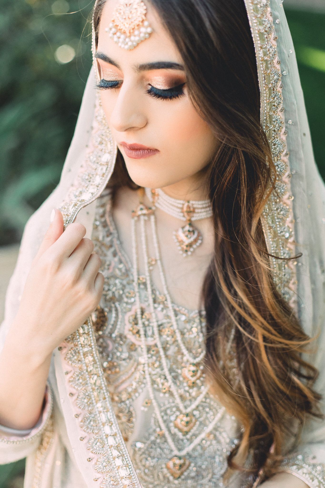 A stunning South Asian bride in ivory embroidered bridal attire with layered pearl jewelry and a delicate dupatta veil.