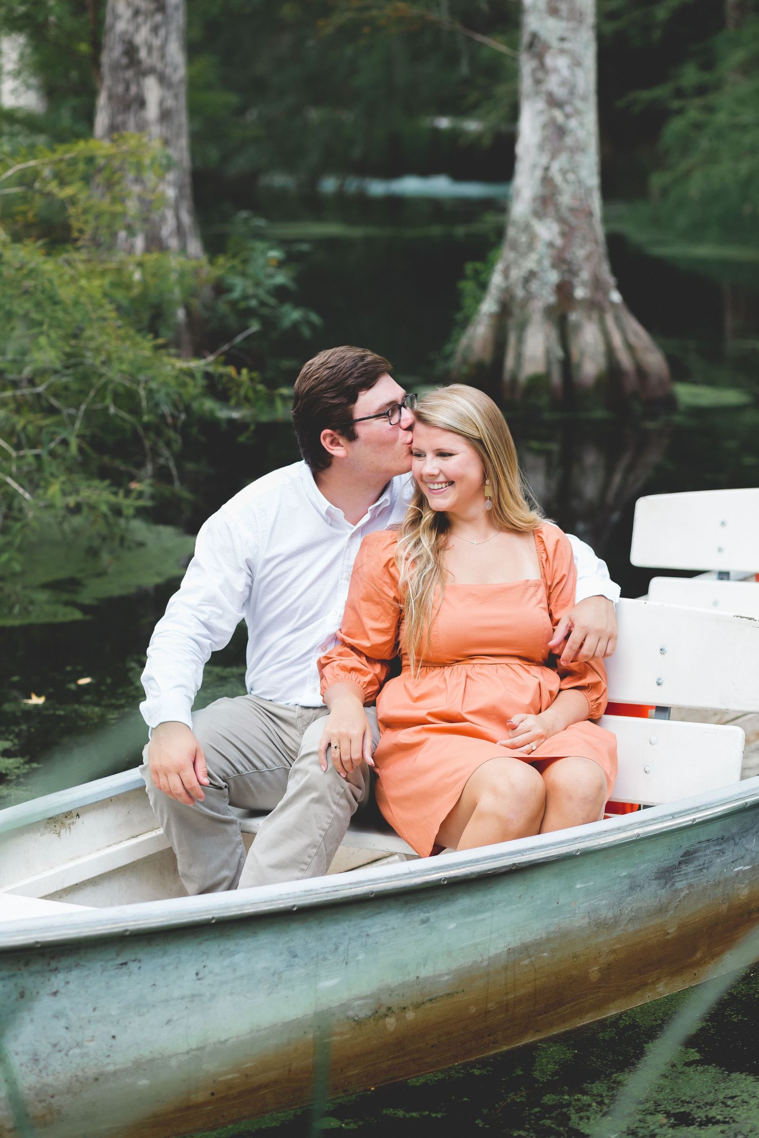 Couple sharing a kiss while sitting in a rowboat surrounded by lush greenery.