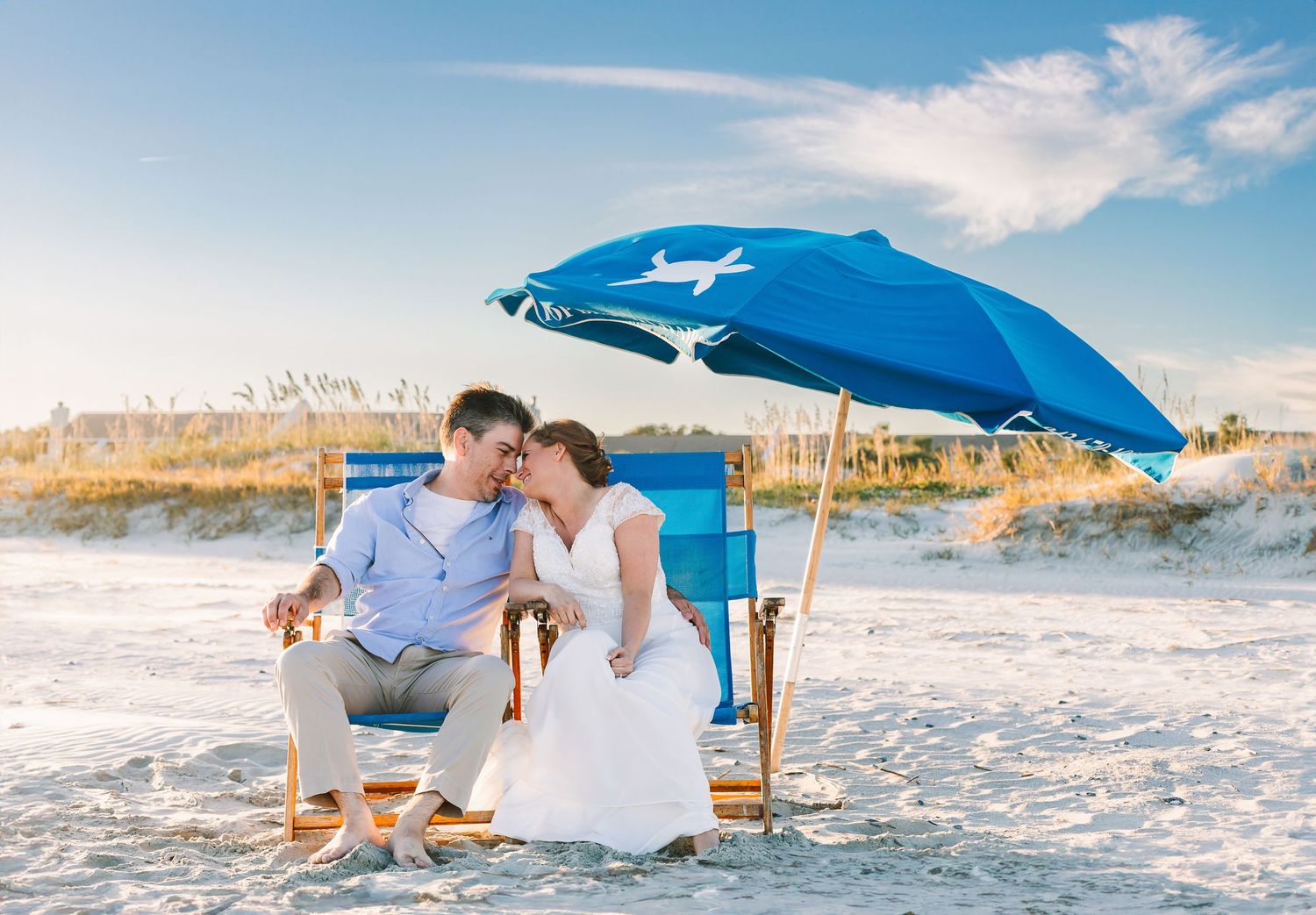 Newlywed couple sitting together under a blue beach umbrella on a sunny sandy beach after their wedding.