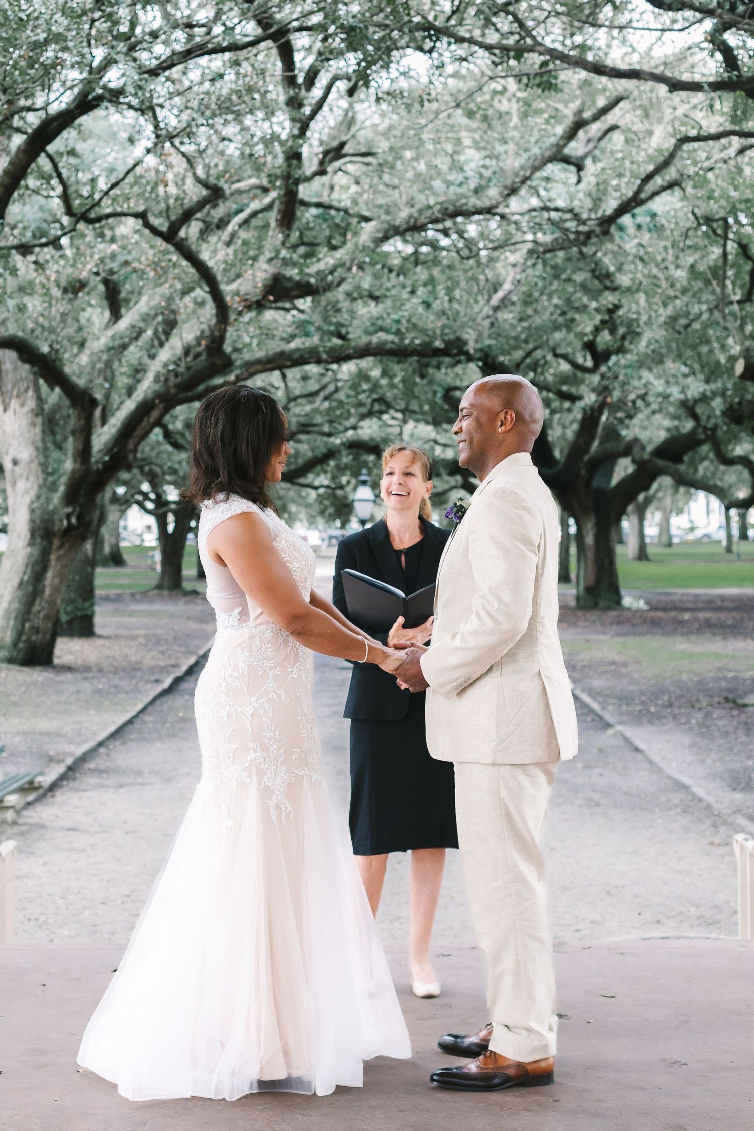 Bride and groom exchange vows outdoors under canopy of oak trees with smiling officiant.