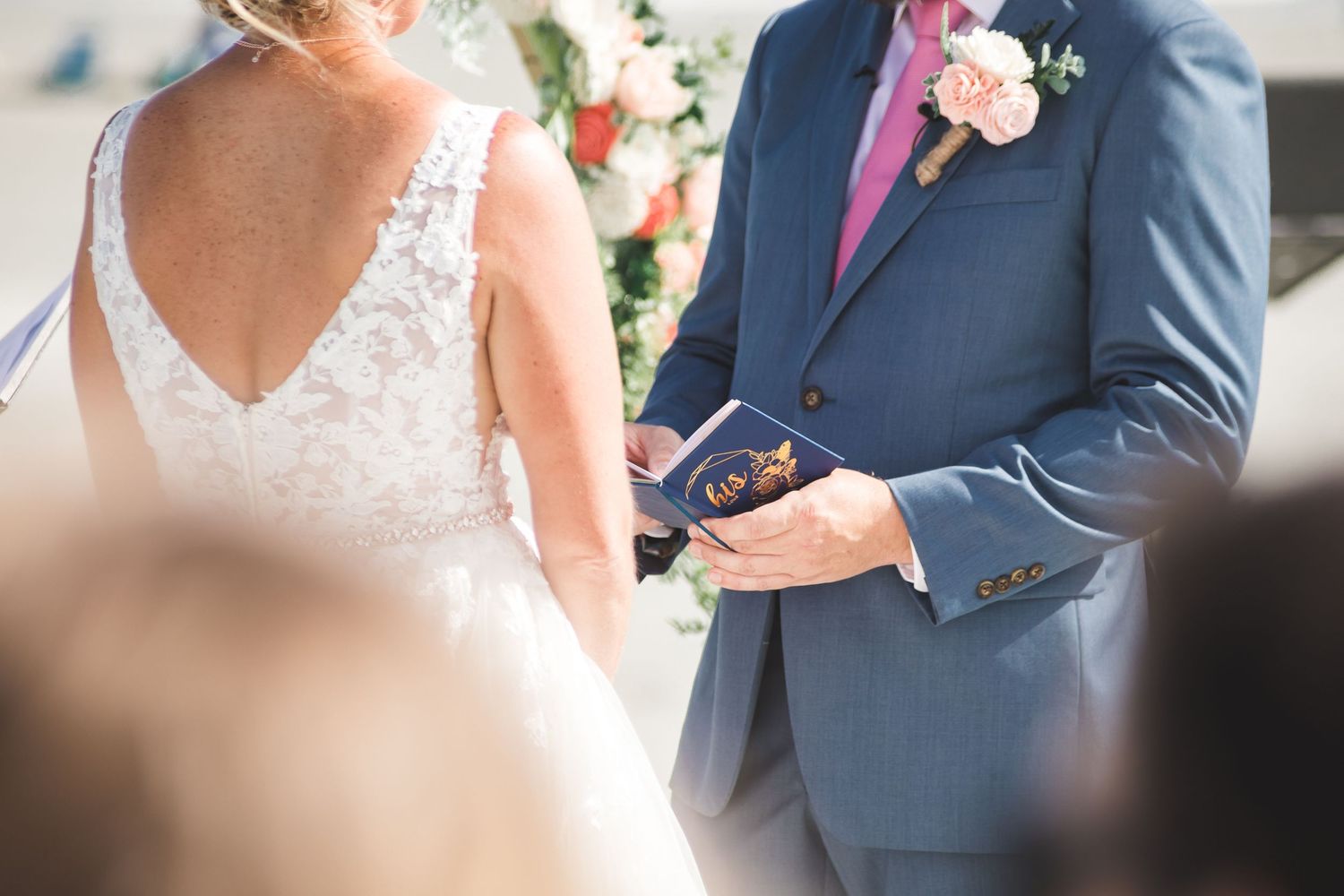 Groom in blue suit holds ring box during outdoor wedding ceremony beside lace-gowned bride.