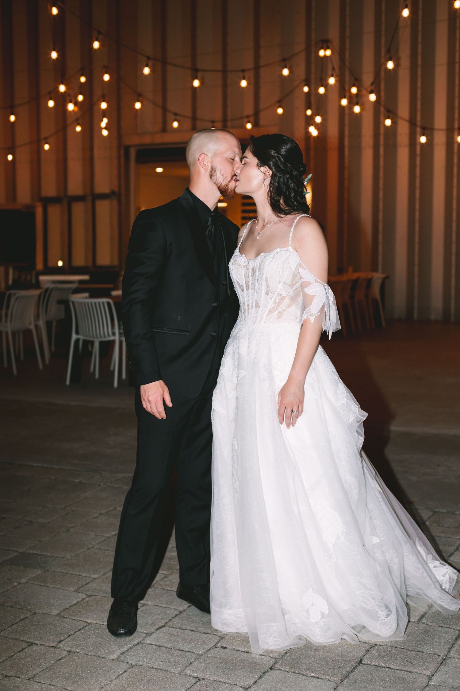 Bride and groom share romantic kiss under string lights at elegant evening wedding venue.