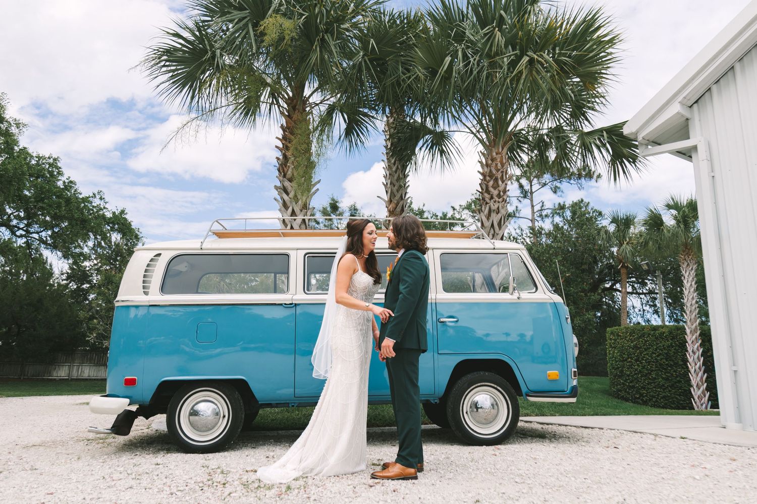 Bride and groom stand before a vintage teal VW bus surrounded by palm trees on their wedding day.