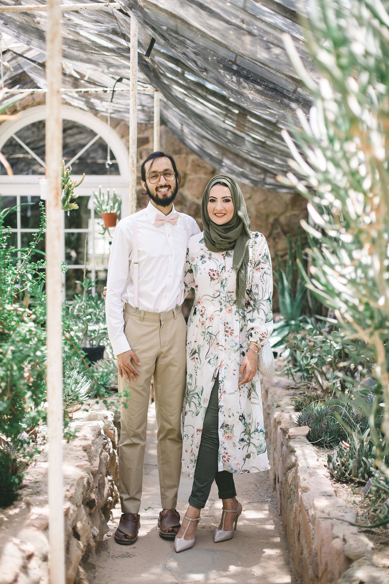 Couple posing in a greenhouse, man in white shirt with bow tie and woman in floral hijab outfit smiling together.