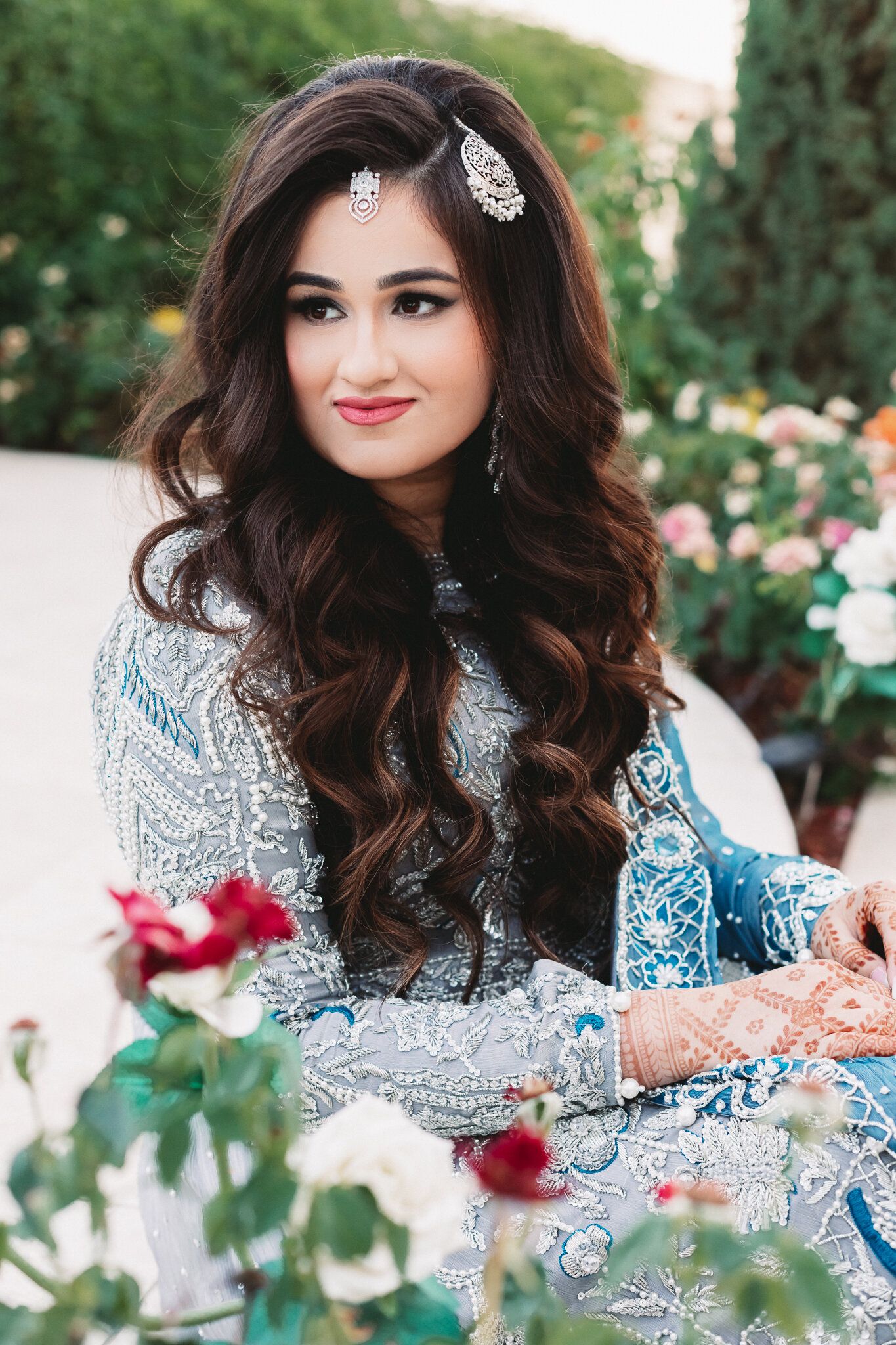 A beautiful South Asian bride in a blue embroidered lehenga with silver jewelry and henna, posing outdoors among roses.