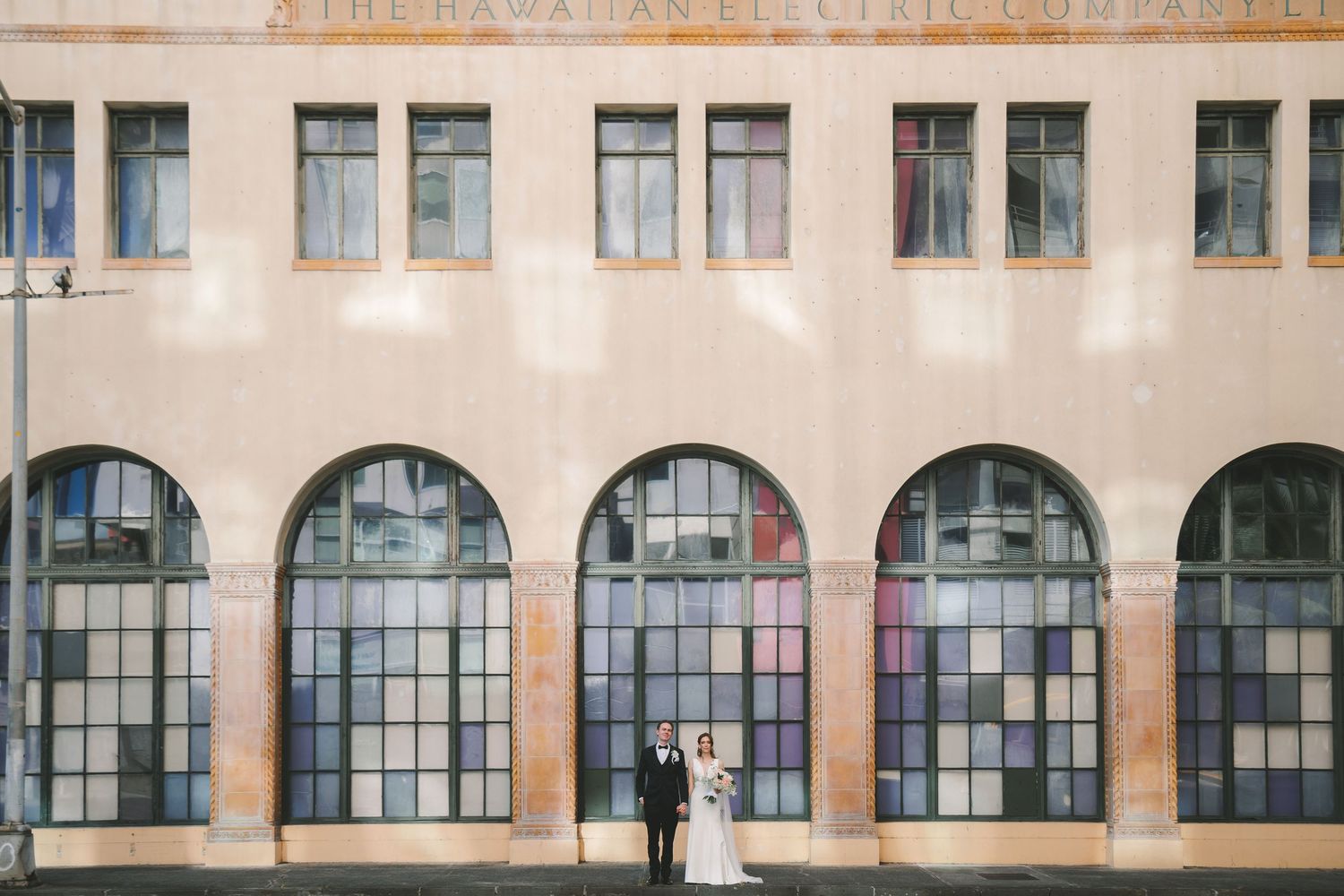 Bride and groom stand before large arched colorful windows of Hawaiian Electric building.