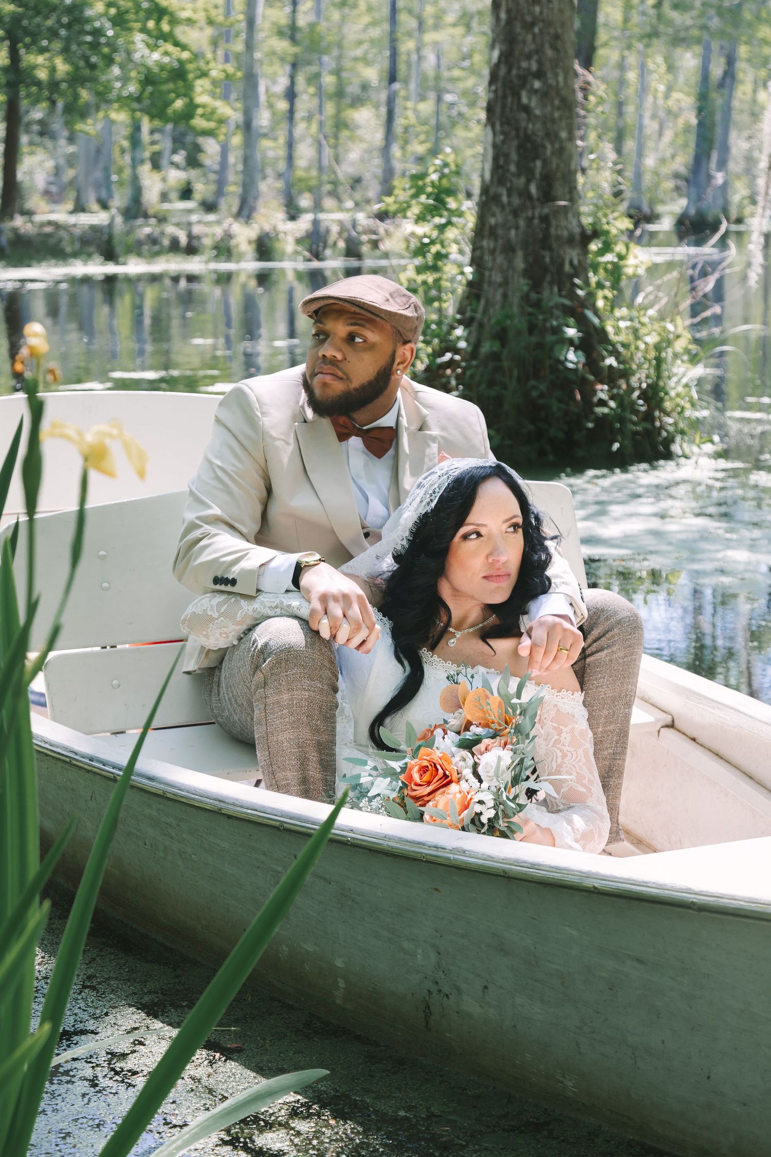 Interracial couple in vintage wedding attire sitting in a white rowboat surrounded by a lush swamp.