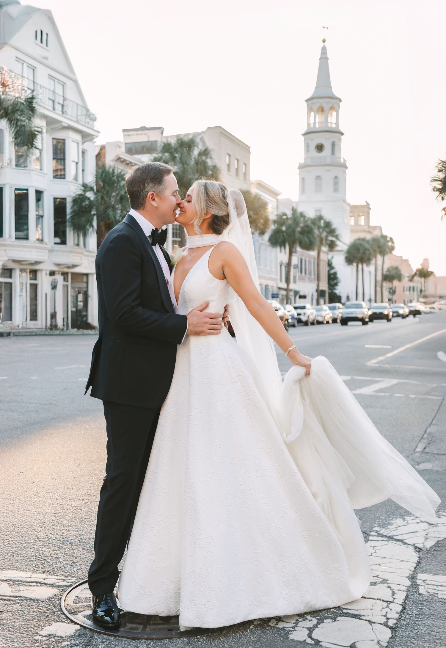 Bride and groom share a romantic kiss on a Charleston street corner with St. Michaels Church steeple at sunset.