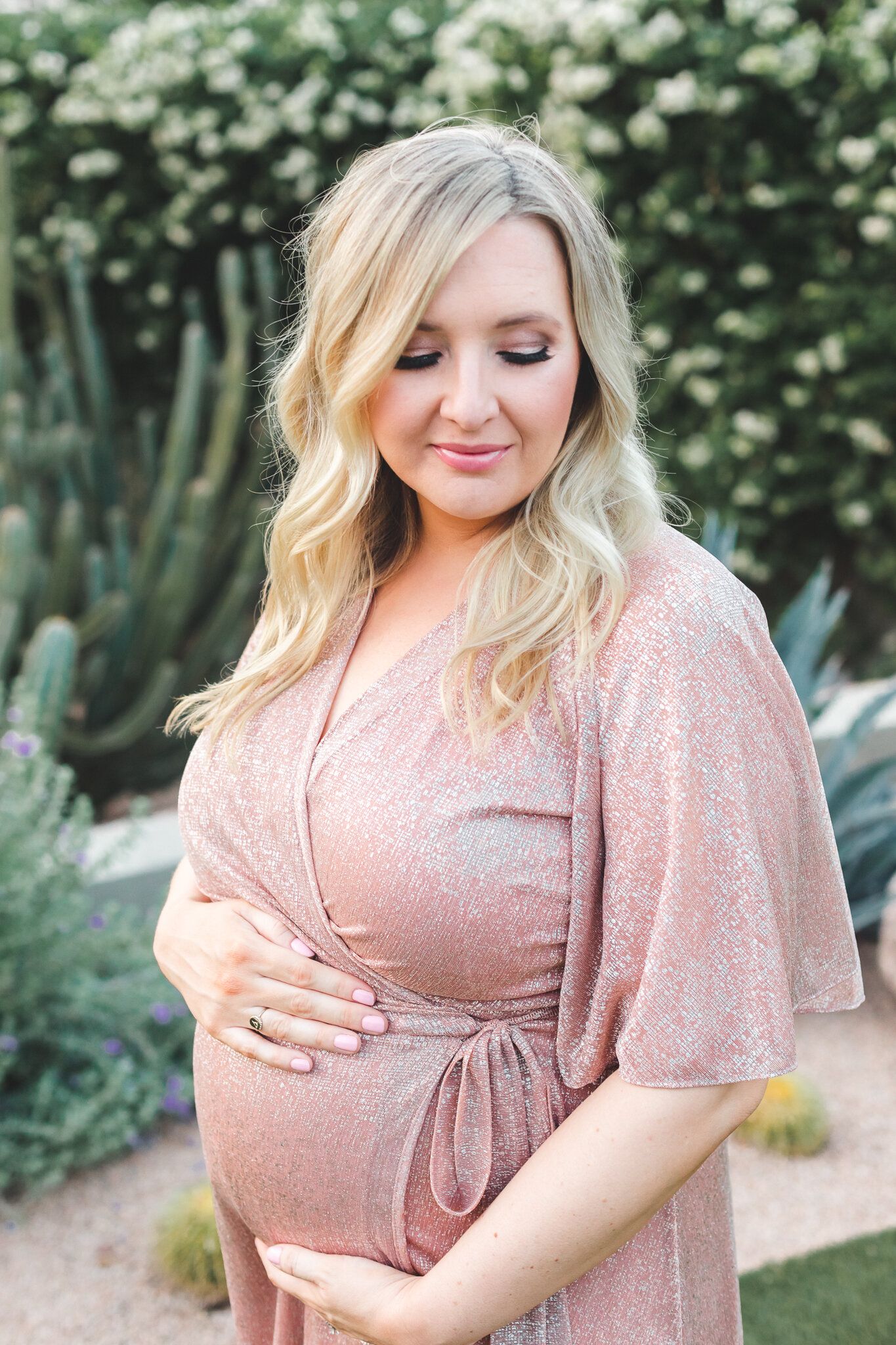 Pregnant woman in pink sequin wrap dress cradling baby bump in cactus garden setting.