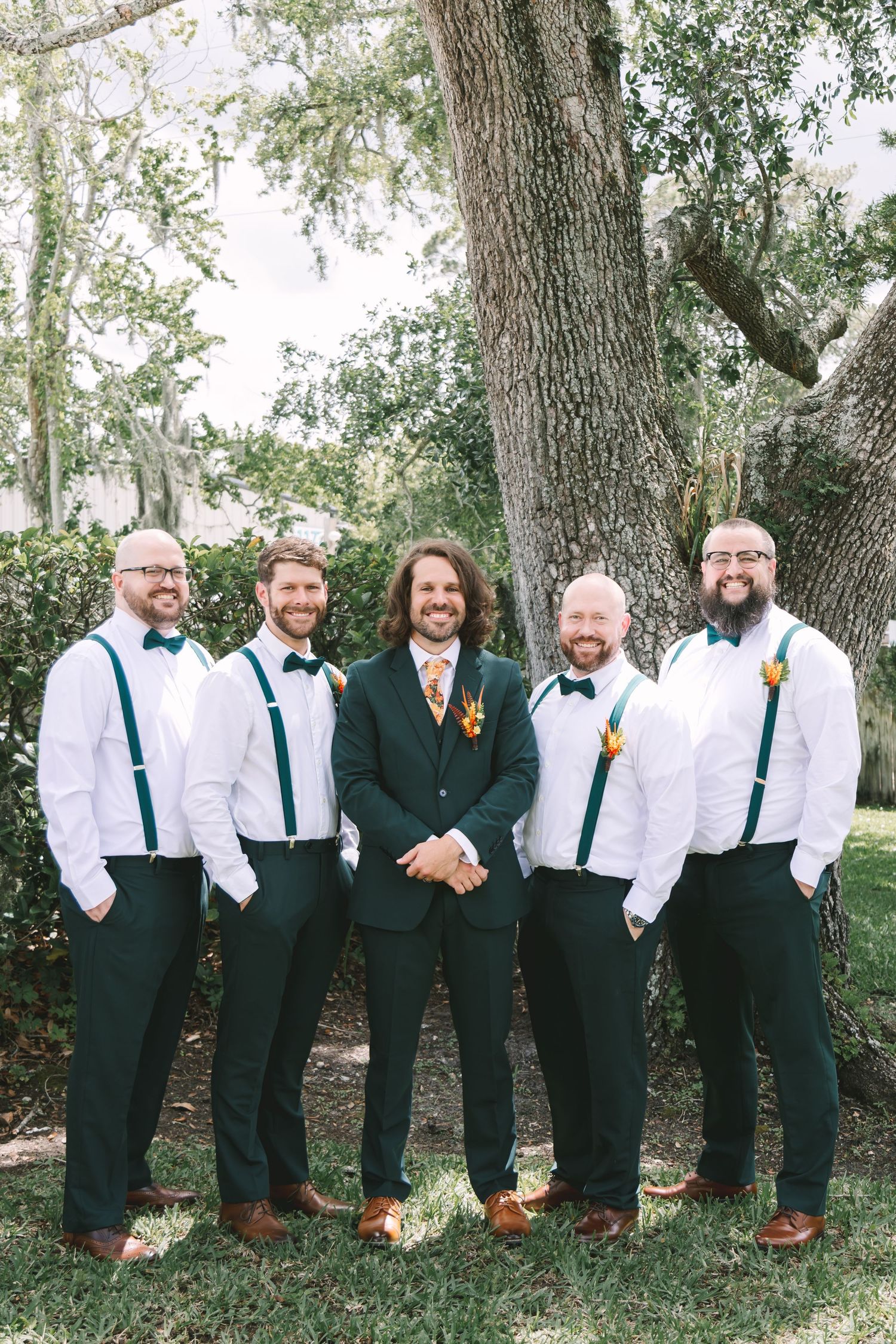 Groom in green suit poses with four groomsmen in white shirts, green suspenders and bow ties outdoors.